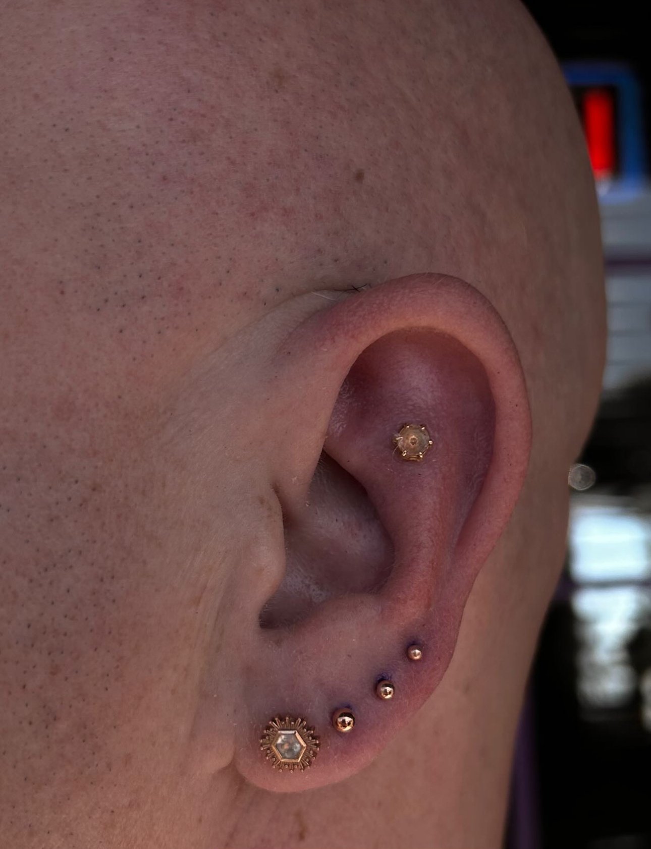 Close-up of a person's ear with multiple piercings, including a helix piercing with gold jewelry, a conch piercing with a gold jewelry, and three earrings in the earlobe, including a hexagonal-shaped earring with a clear stone.