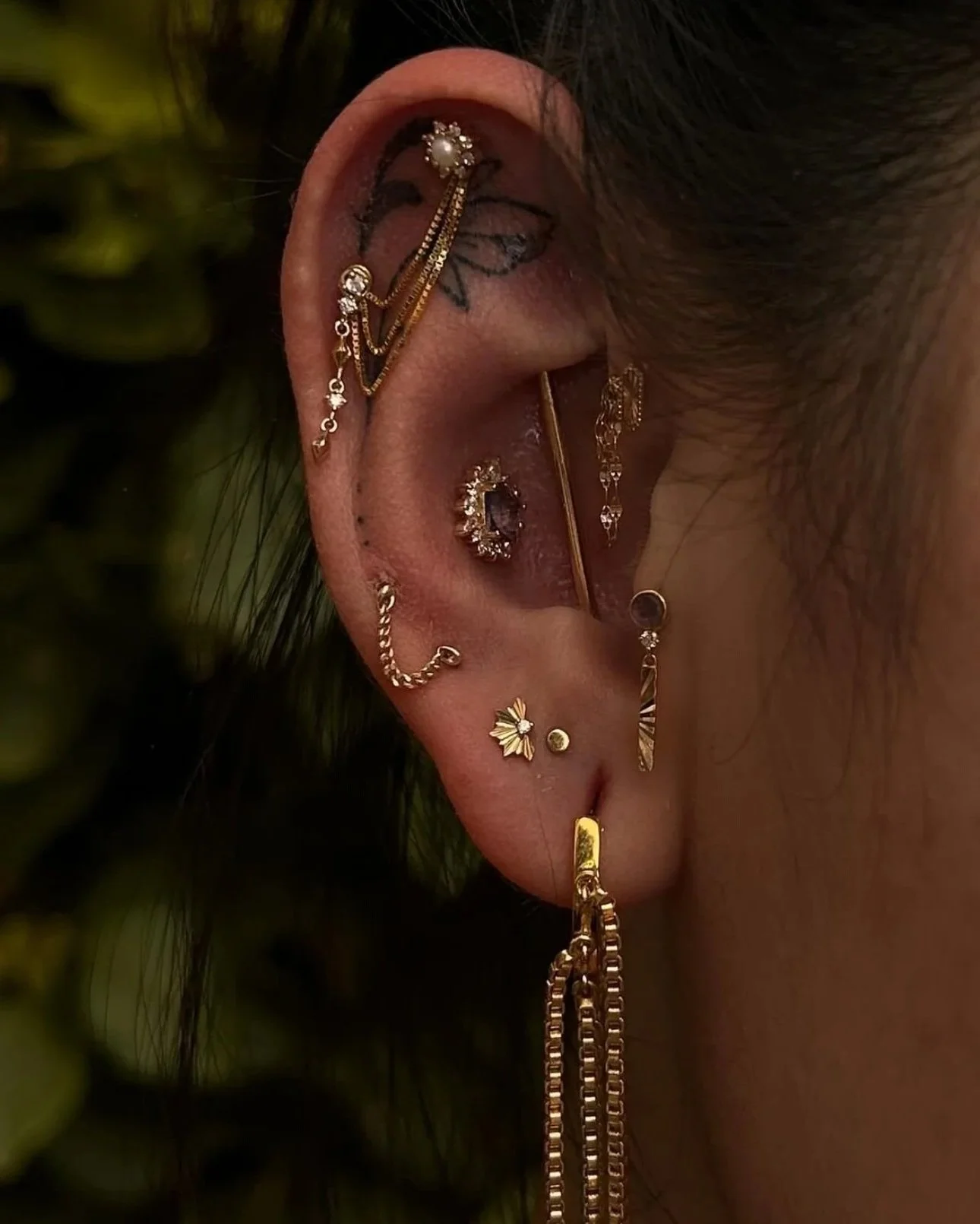 Close-up of a person's ear adorned with multiple gold and silver earrings, including chains, studs, and hoops, with a black tattoo behind the ear and dark hair partially covering it.