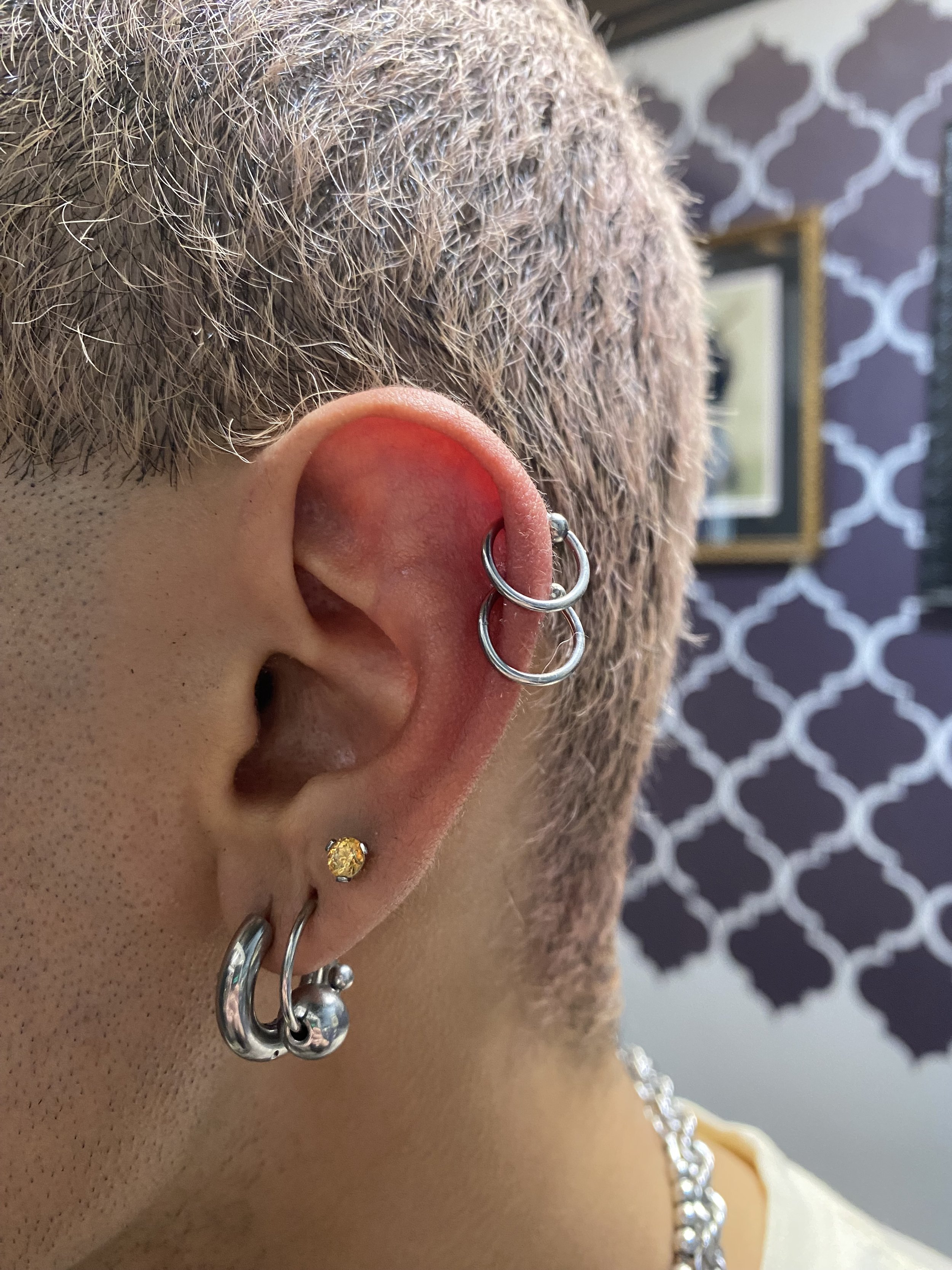 Close-up of a person's ear with multiple earrings, including a multi-loop silver hoop earring, a small gold stud earring, and a small silver hoop earring, set against a background with a dark patterned wall and a picture frame.