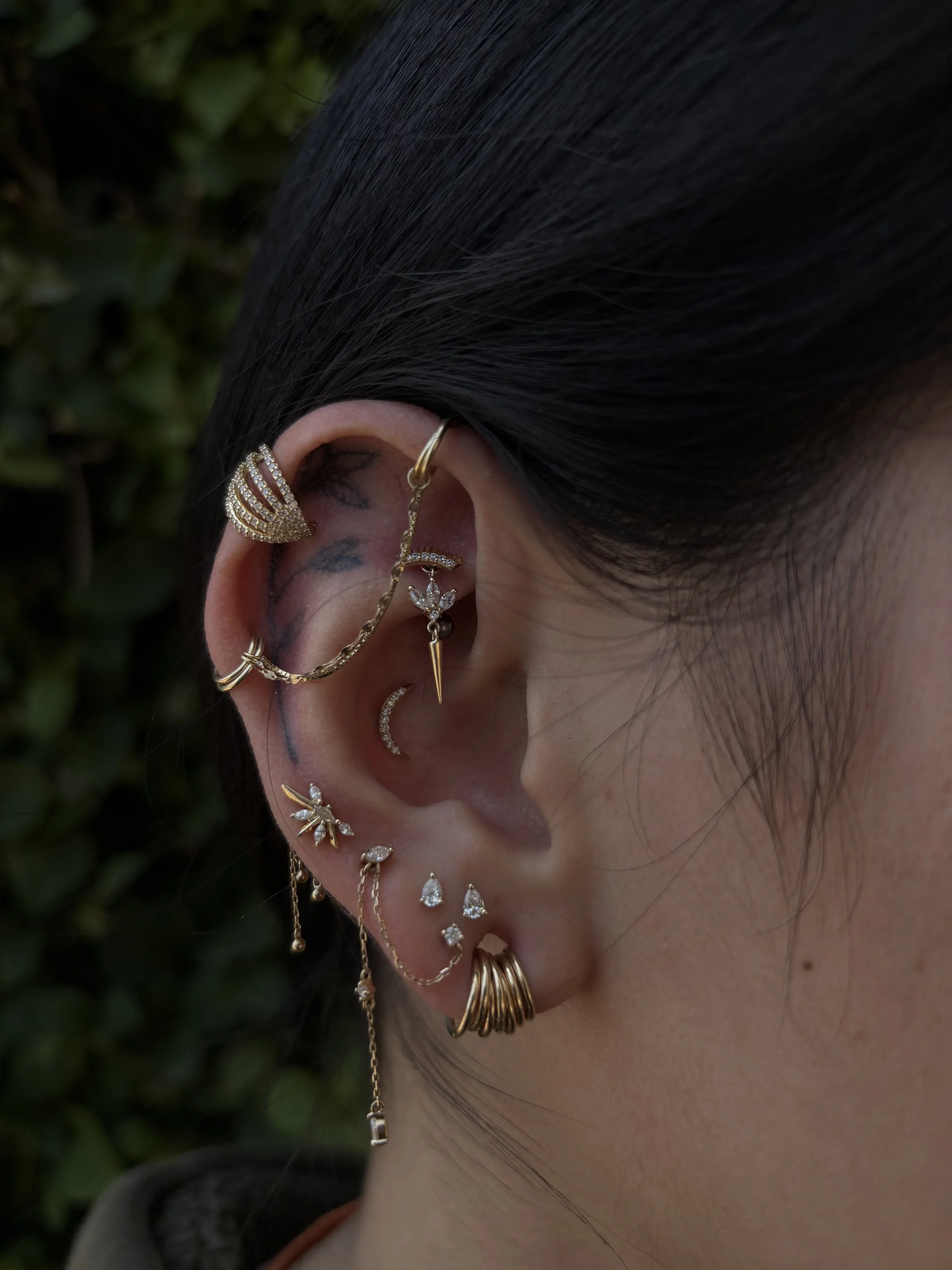 Close-up of a person's ear and multiple gold earrings with various designs and embedded stones, with black hair covering part of the face and blurred green foliage in the background.
