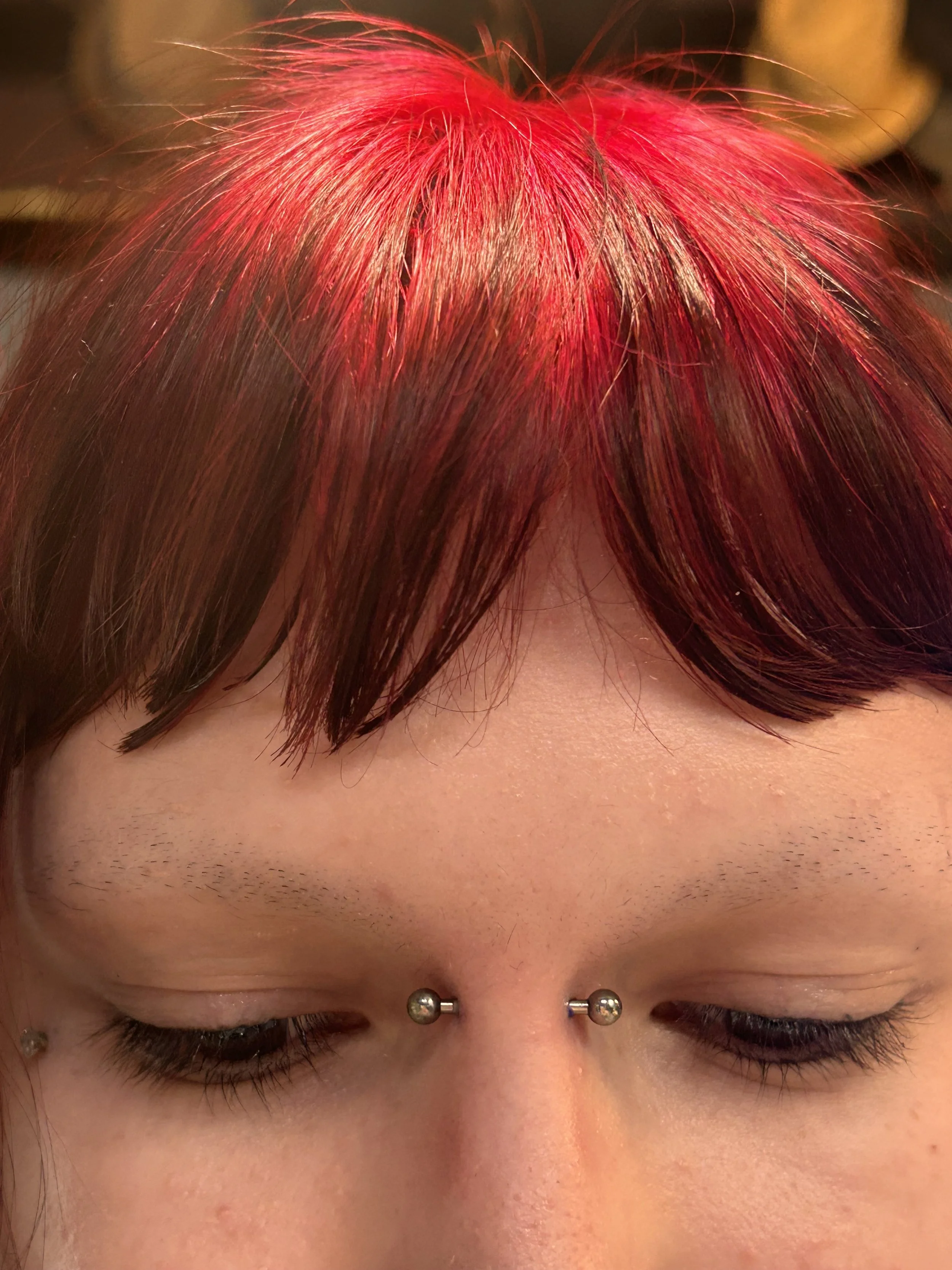 Close-up of a person's forehead and closed eyes, with red hair covering the forehead and a unique eyebrow piercing. The person also has a nose piercing with silver studs.