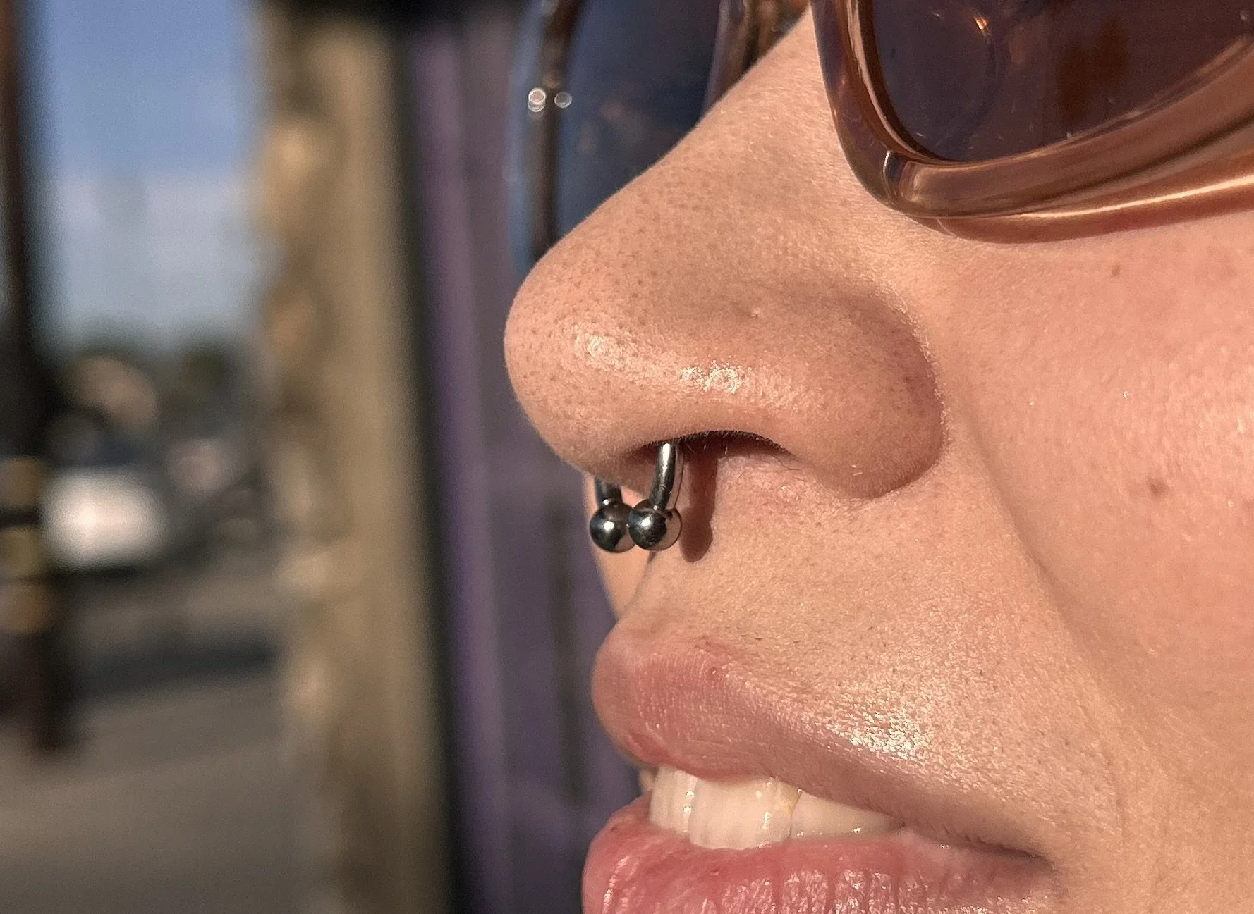 Close-up of a person's nose with a septum piercing, wearing sunglasses, with a blurred outdoor background.