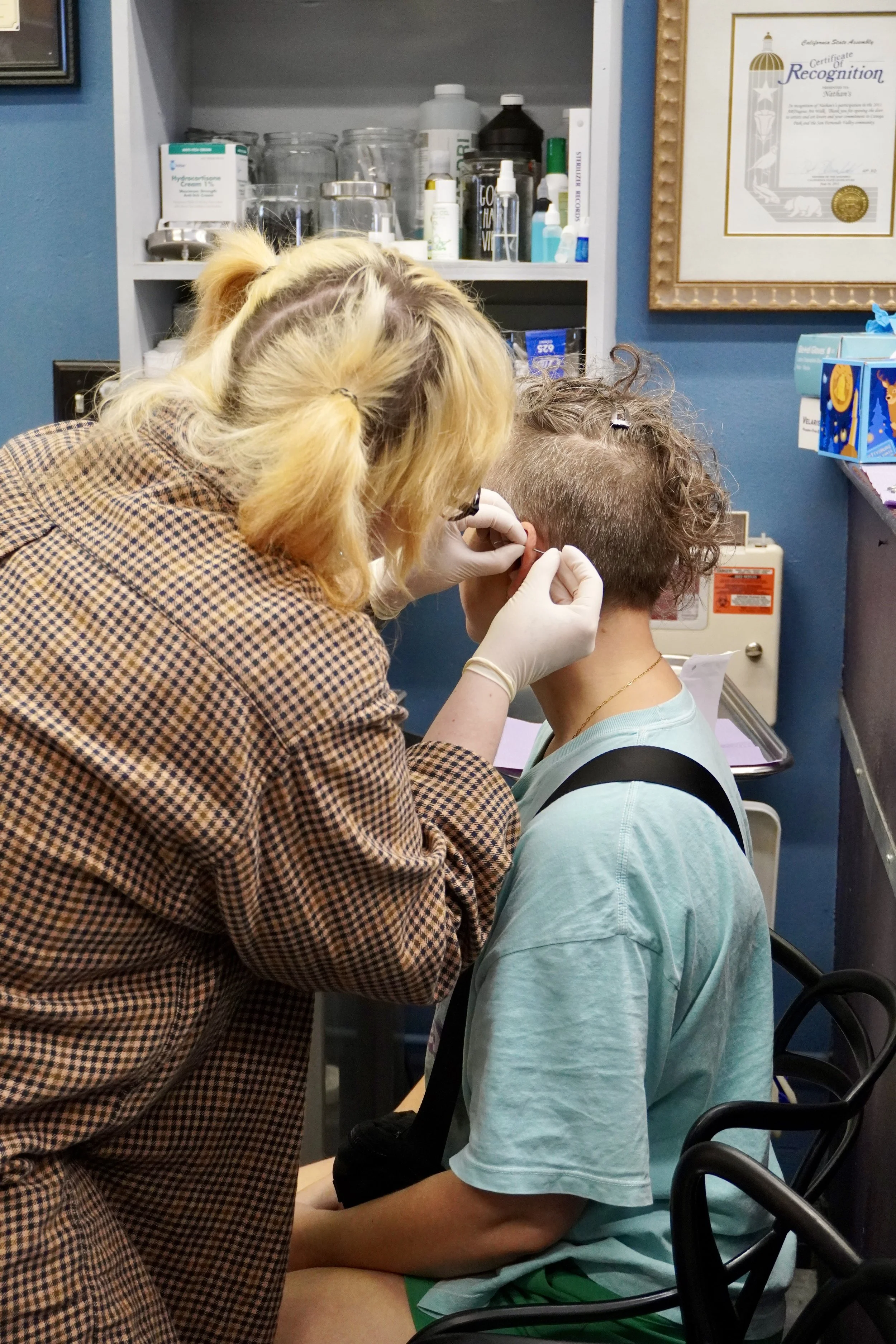 A person with blonde hair in pigtails is giving a young boy a vaccination or shot in an examination room. The boy is wearing a light blue t-shirt and green shorts and is sitting in a chair with black armrests. The room has open shelves with medical supplies and a framed certificate on the wall.