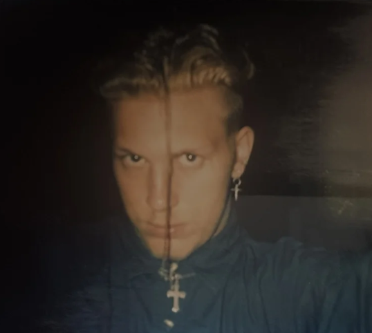 A young man with short, styled hair wearing a dark jacket with a zipper and a cross pendant necklace, taking a selfie against a dark background.
