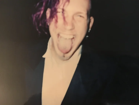 Person with pink and purple hair sticking out their tongue and smiling, wearing a dark jacket and white shirt.