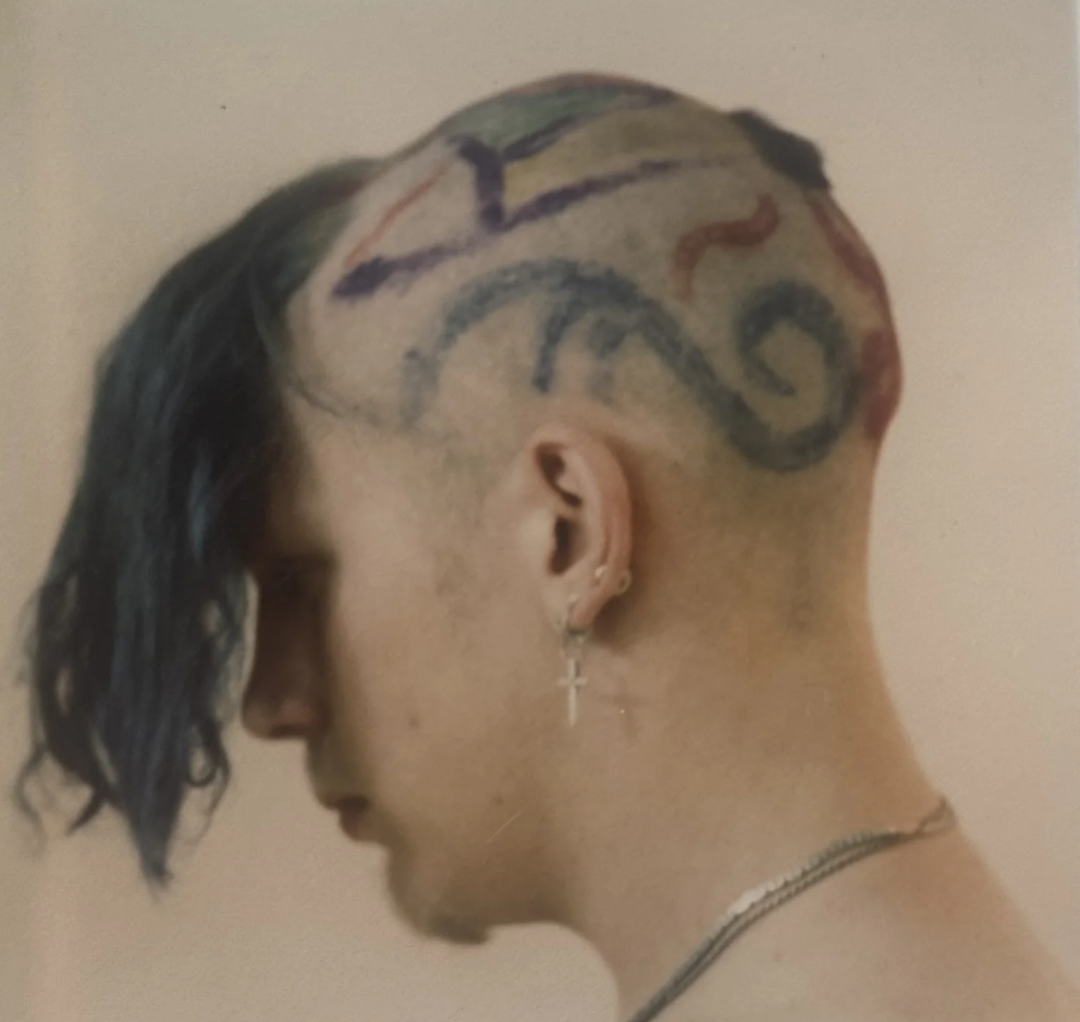 Person with a mohawk hairstyle featuring a colorful tattoo design on the shaved side of the head, wearing earrings and a necklace, with their head tilted downward.