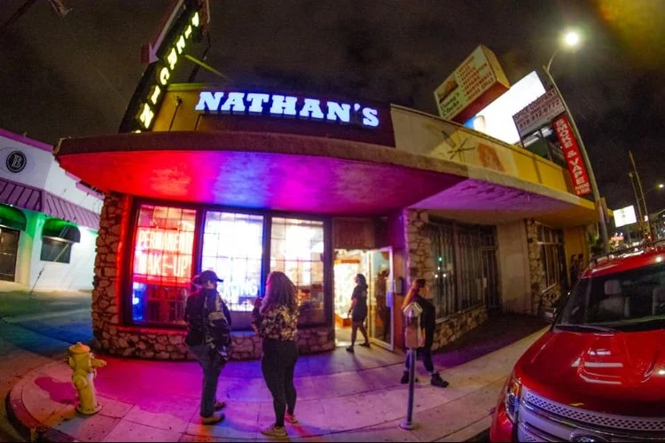Night scene outside Nathan's restaurant with people waiting in line, illuminated signs, and a red car parked nearby.