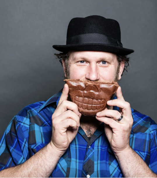 Man wearing a black fedora and blue plaid shirt holding a chocolate skull mask in front of his face.