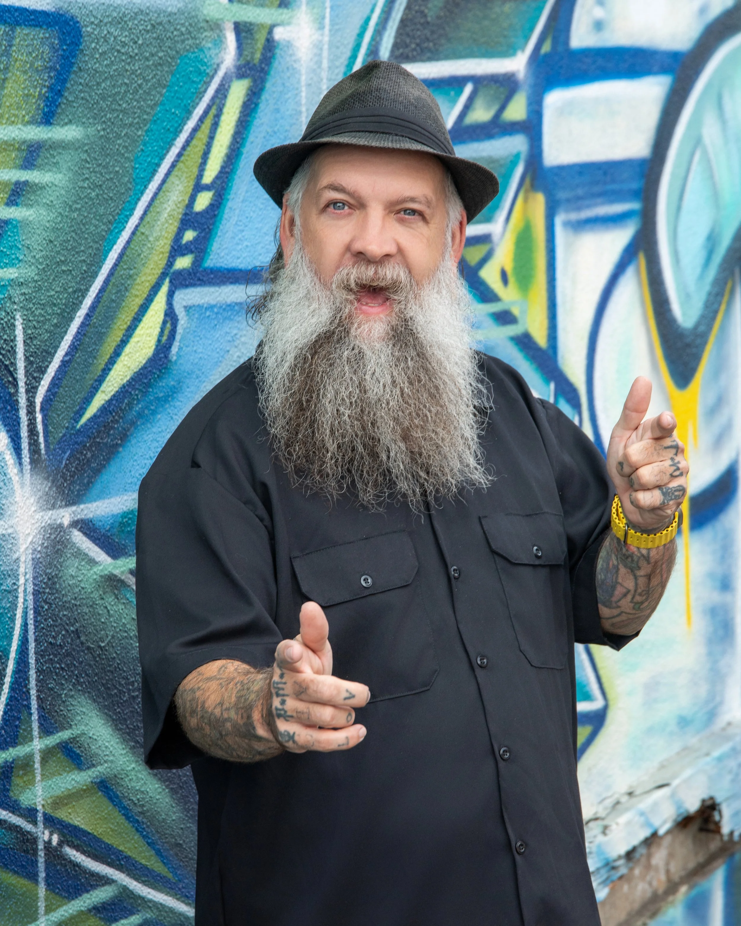 A man with a long gray beard and tattoos, wearing a black shirt, a gray fedora, and a yellow watch, is standing in front of a colorful graffiti wall with blue, green, yellow, and black designs.