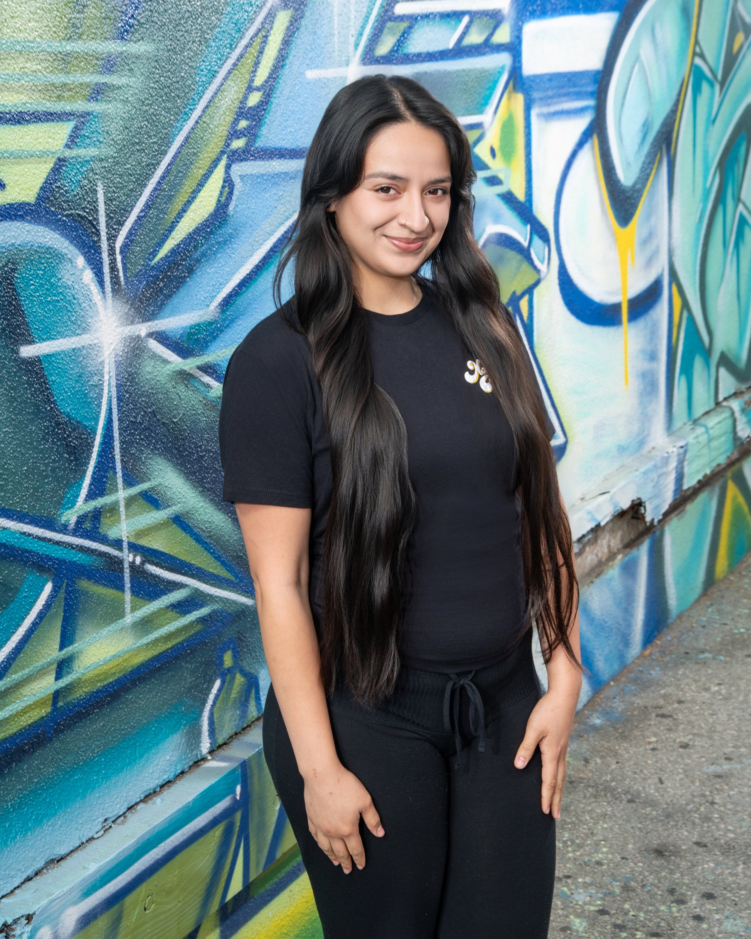 A young woman with long dark hair standing against a colorful graffiti wall, wearing a black t-shirt and black pants, smiling at the camera.
