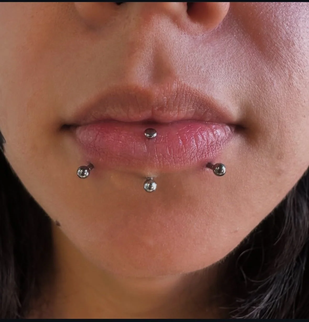 Close-up of a person's lips with four lip piercings, two on the lower lip and two on the upper lip, with a focus on the pink, slightly glossy lips and surrounding skin.