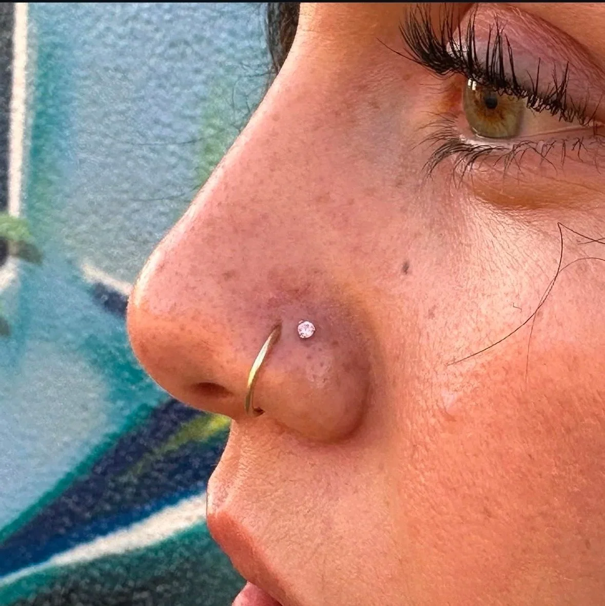 Close-up of a person's face showing a nose piercing with a small gemstone and a gold hoop, part of an eye with brown iris, and skin with freckles and a mole.