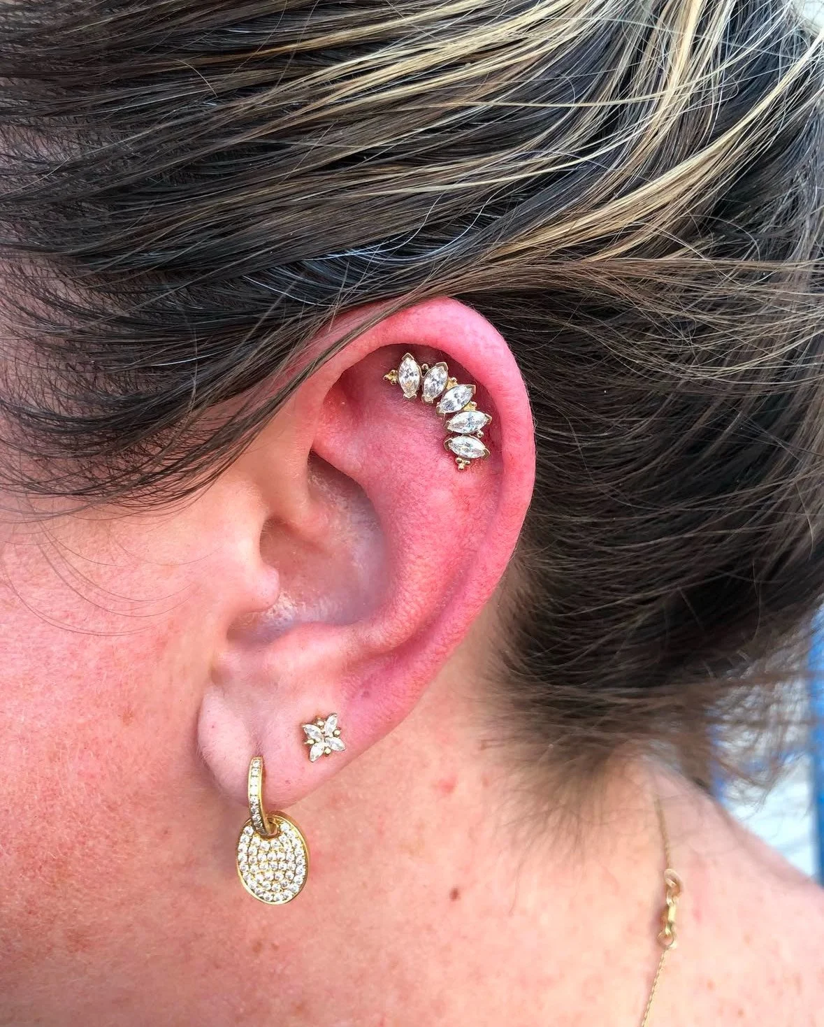 Close-up of a person's ear with multiple earrings: a diamond-studded hoop, a butterfly-shaped stud, and a marquise-shaped ear cuff with several gemstones.