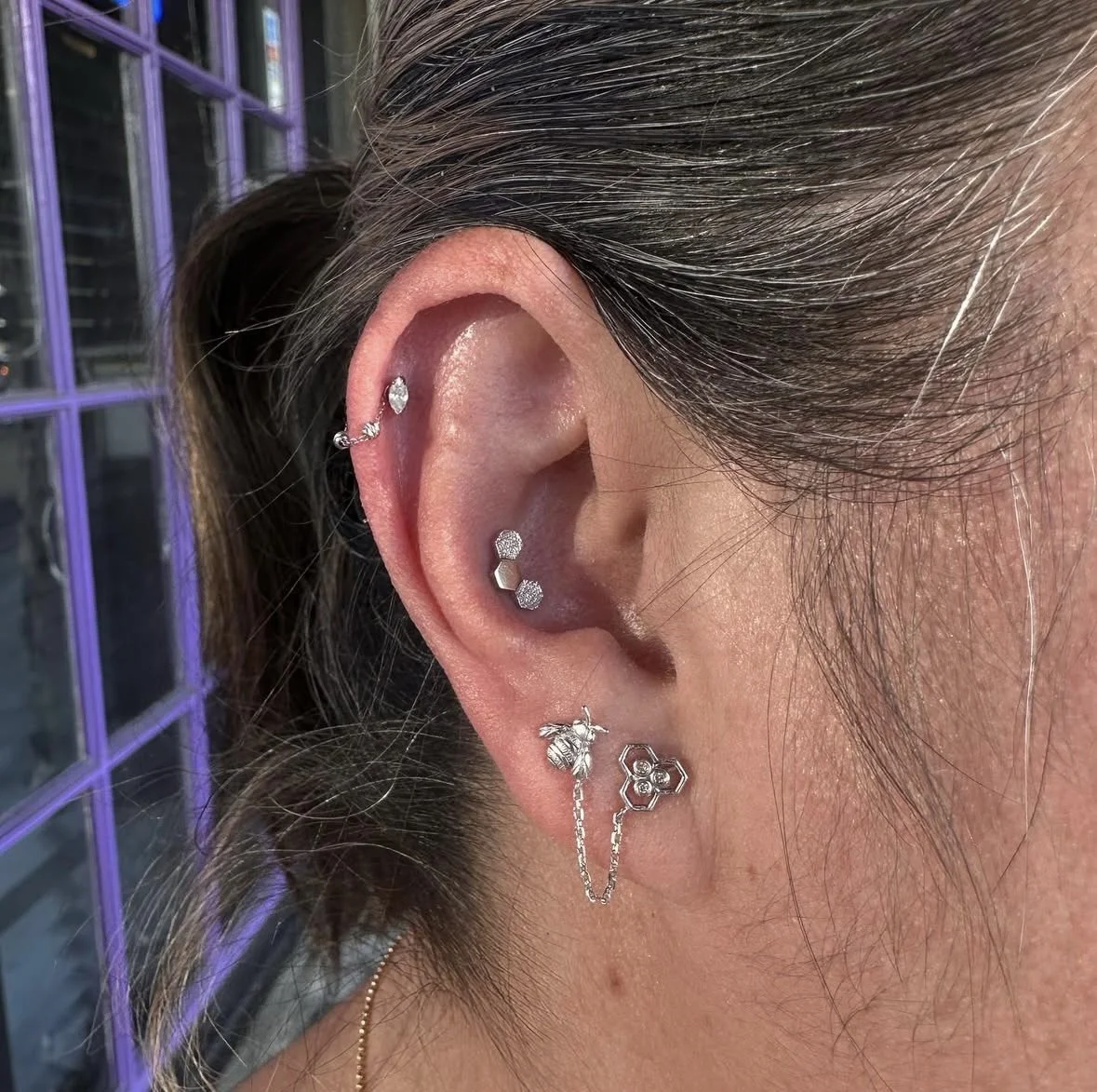 Close-up of a person's ear with multiple piercings, showcasing various silver earrings including studs, hoops, and chains with geometric and textured designs.