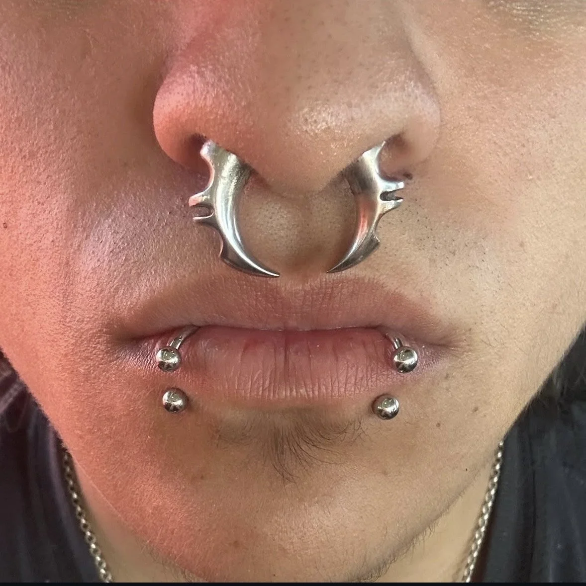 Close-up of a person's face with multiple facial piercings including a septum piercing with a chrome ring, four lip piercings with small ball studs, and a chain necklace.