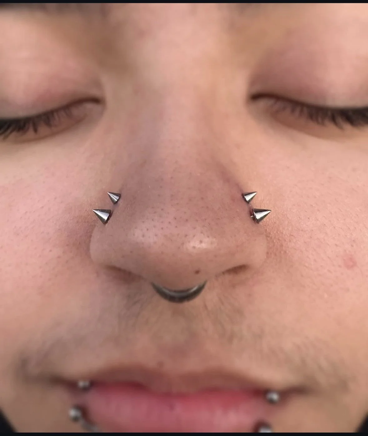 Close-up of a person's face with closed eyes, multiple facial piercings including a septum piercing, and black lip rings.