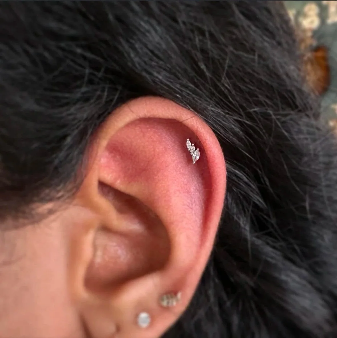 Close-up of a person's ear with three small earrings, one with a leaf-shaped diamond and two smaller round diamond studs, surrounded by dark hair.