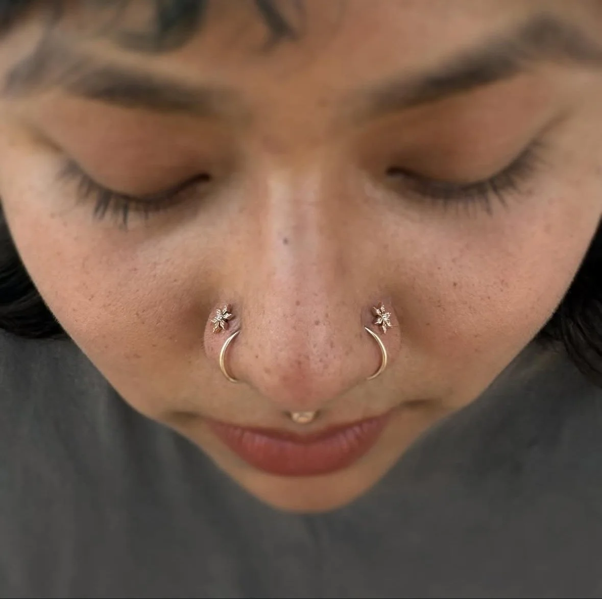 Close-up of a woman's face with closed eyes, showcasing multiple facial piercings including nose hoops and a septum piercing, with natural makeup and freckles.