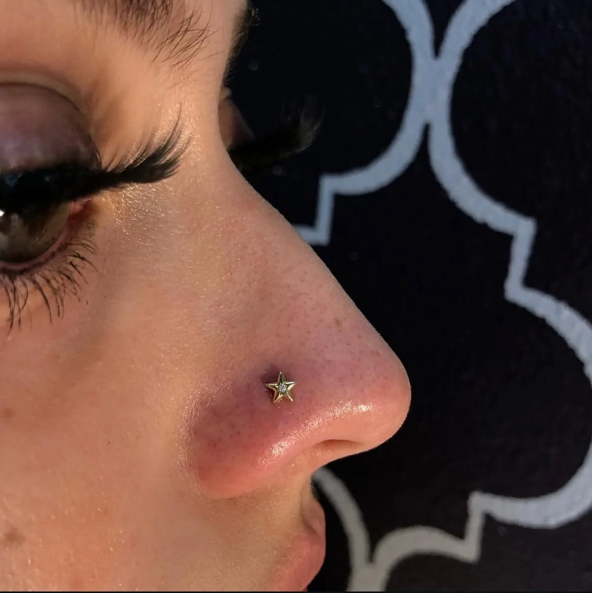 Close-up of a woman's face showing her nose with a gold star-shaped nose ring, part of her eye, and long eyelashes, with a black and white patterned background.