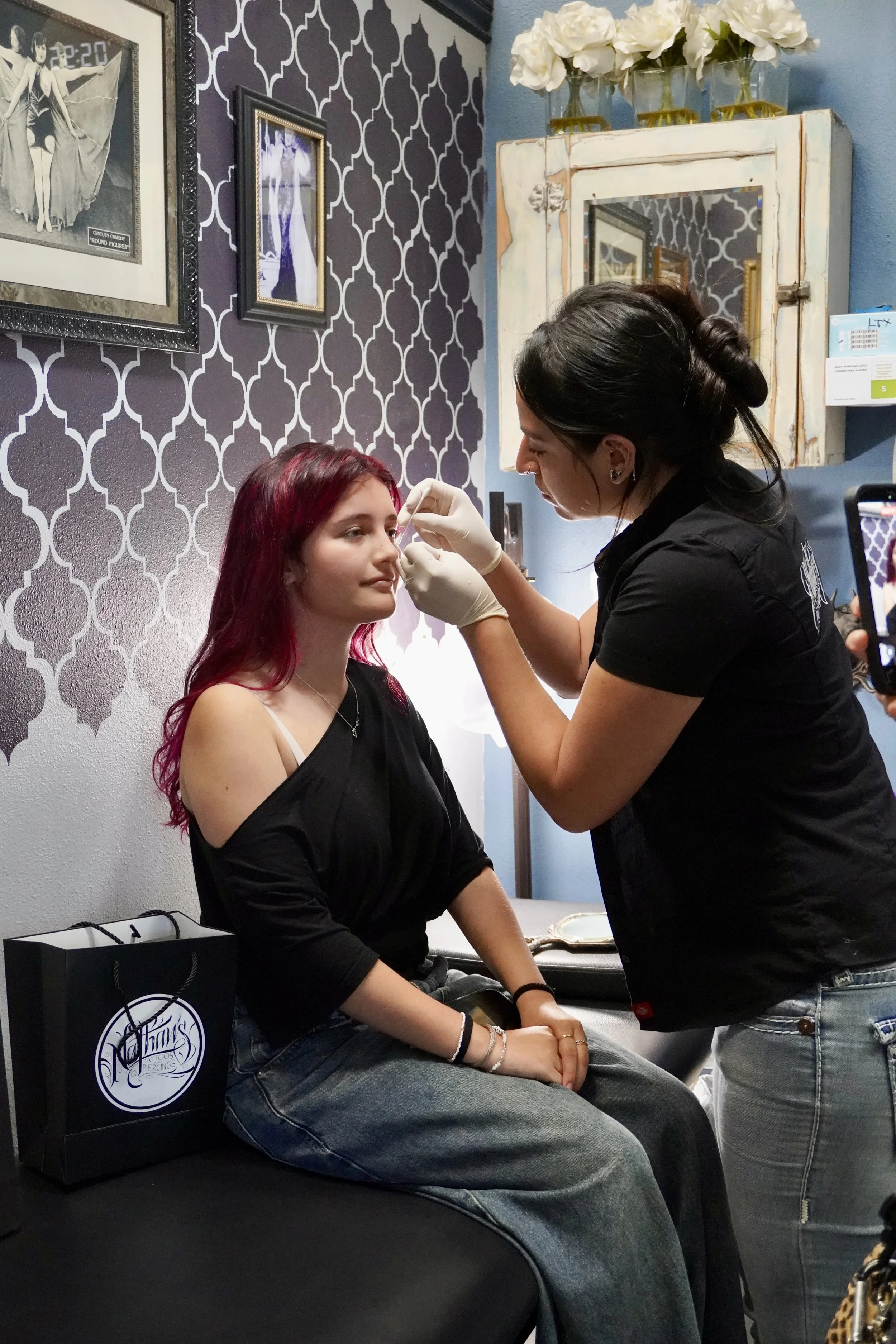 A woman with long reddish hair sitting on a black surface, getting a nose piercing from a piercing artist who has dark hair in a bun. The woman is wearing a black off-shoulder top and jeans. There are framed pictures on the patterned wall behind them and a white distressed cabinet above them with white flowers in glass vases. There is also a black shopping bag with a white logo on it next to the woman.