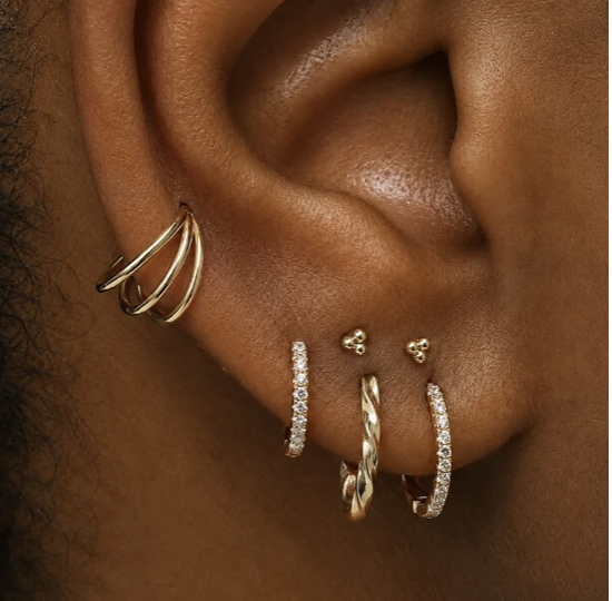 Close-up of a person's ear with multiple gold earrings, including a triple hoop, a small stud, a twisted hoop, and a bejeweled hoop, set against brown skin.