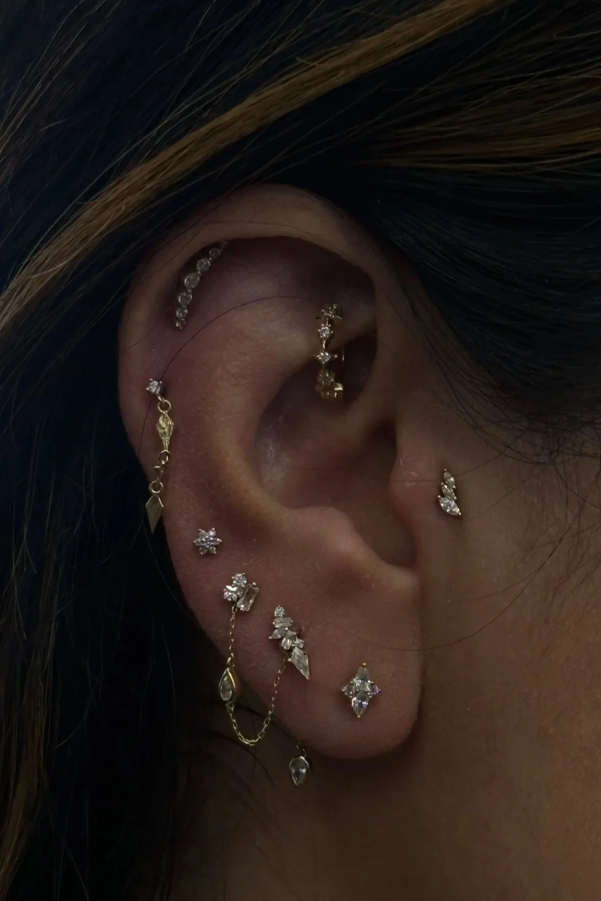 Close-up of a person's ear adorned with multiple gold and diamond jewelry pieces, including earrings, a chain, and ear cuffs.