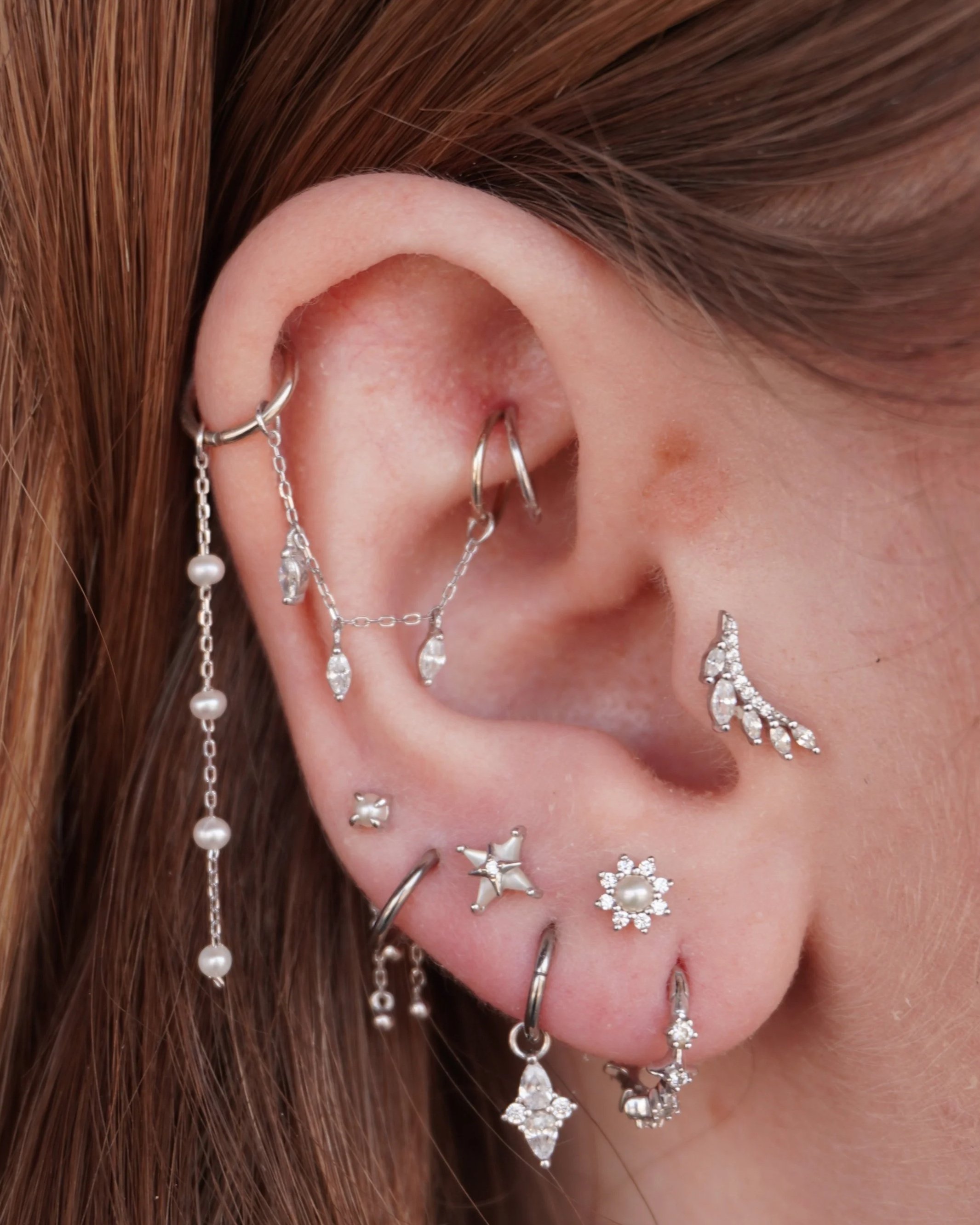 Close-up of a person's ear adorned with multiple silver earrings and jewelry, surrounded by reddish-brown hair.