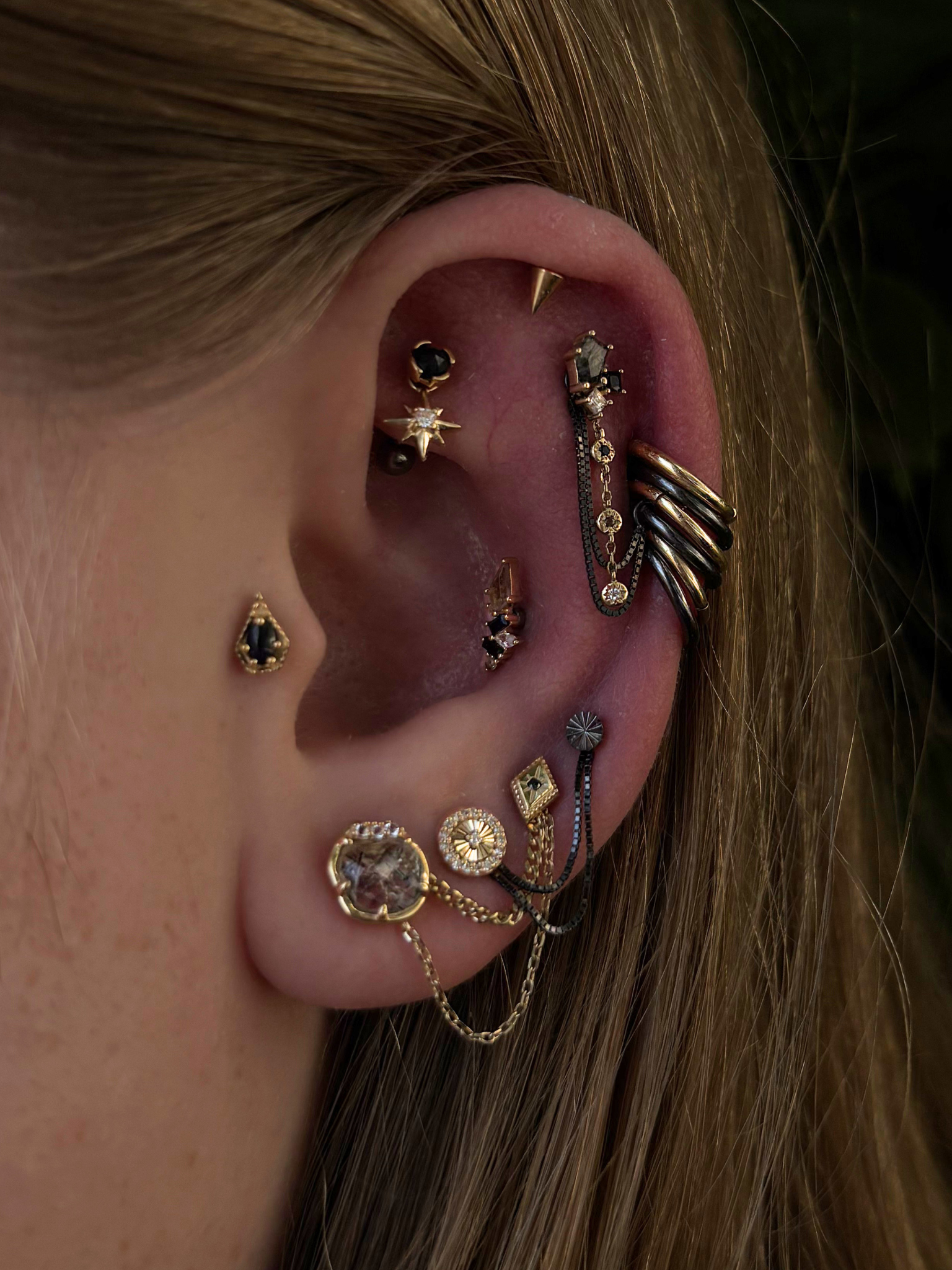Close-up of a human ear adorned with multiple earrings and jewelry, including gold hoops, chains, and various decorative piercings, with long, light brown hair surrounding the ear.