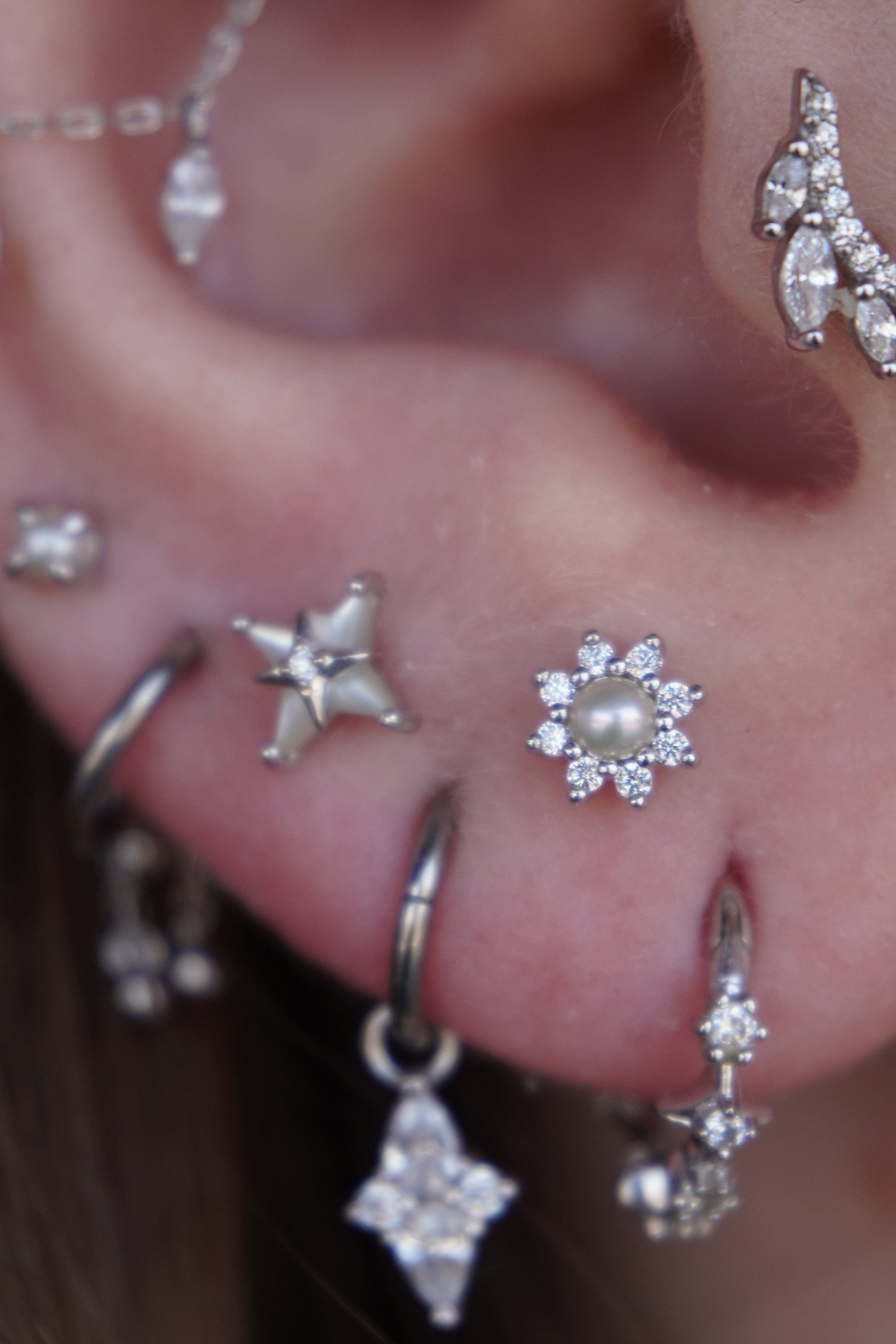 Close-up of a person's ear adorned with multiple white gold and diamond earrings, including a star-shaped earring with a large central gem, a snowflake-shaped earring with a pearl in the center, and a layered teardrop-shaped earring with diamonds.