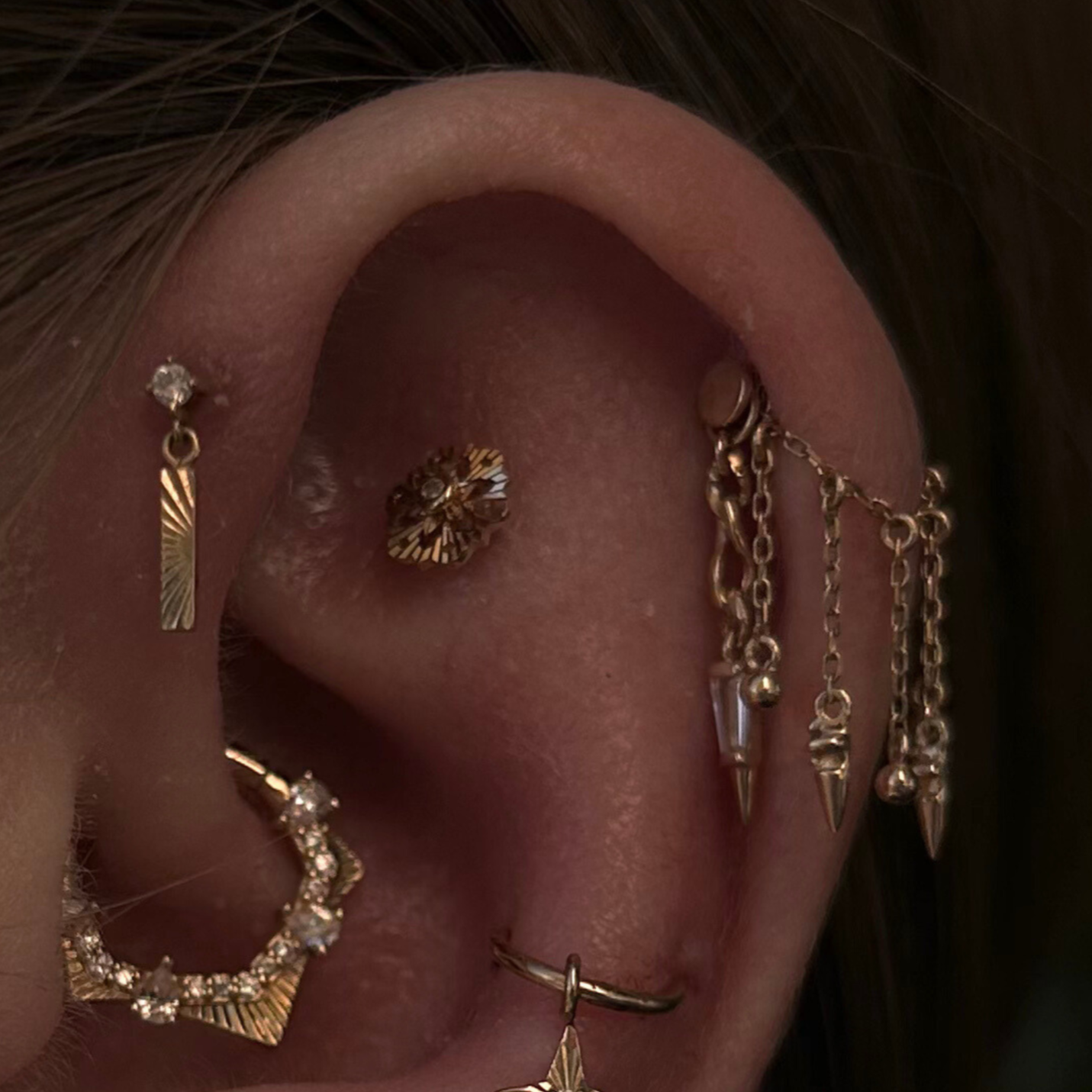 Close-up of a person's ear decorated with multiple gold earrings, including a small diamond stud, a diagonal gold bar earring, a flower-shaped earring, and several chain earrings with pointed details.