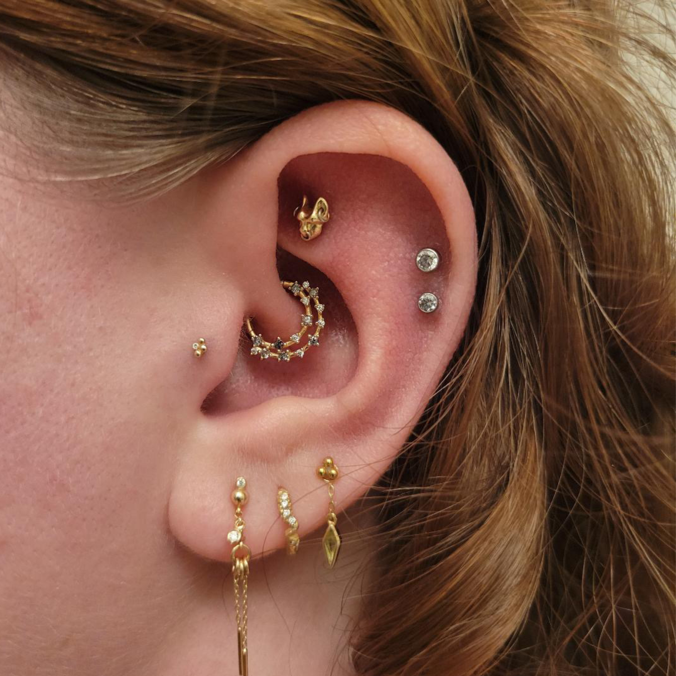 Close-up of a person's ear with multiple gold earrings including a hoop, a stud, and dangling jewelry, with brown hair parting visible.