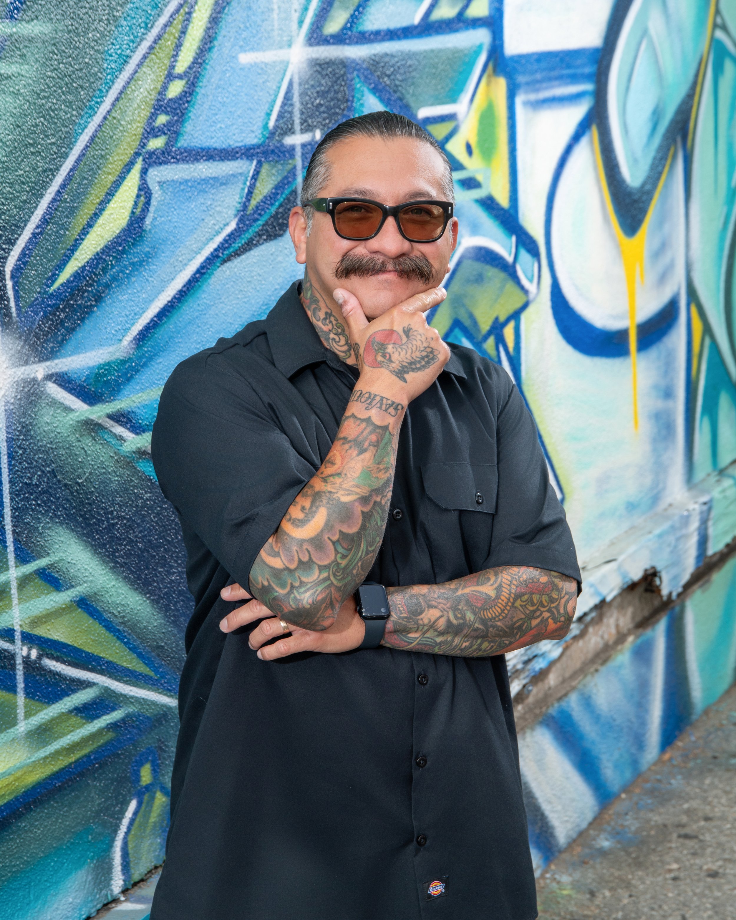 A man with sunglasses and tattoos on his arms and neck, wearing a black Dickies shirt, standing with his hand on his chin in front of a colorful graffiti wall.