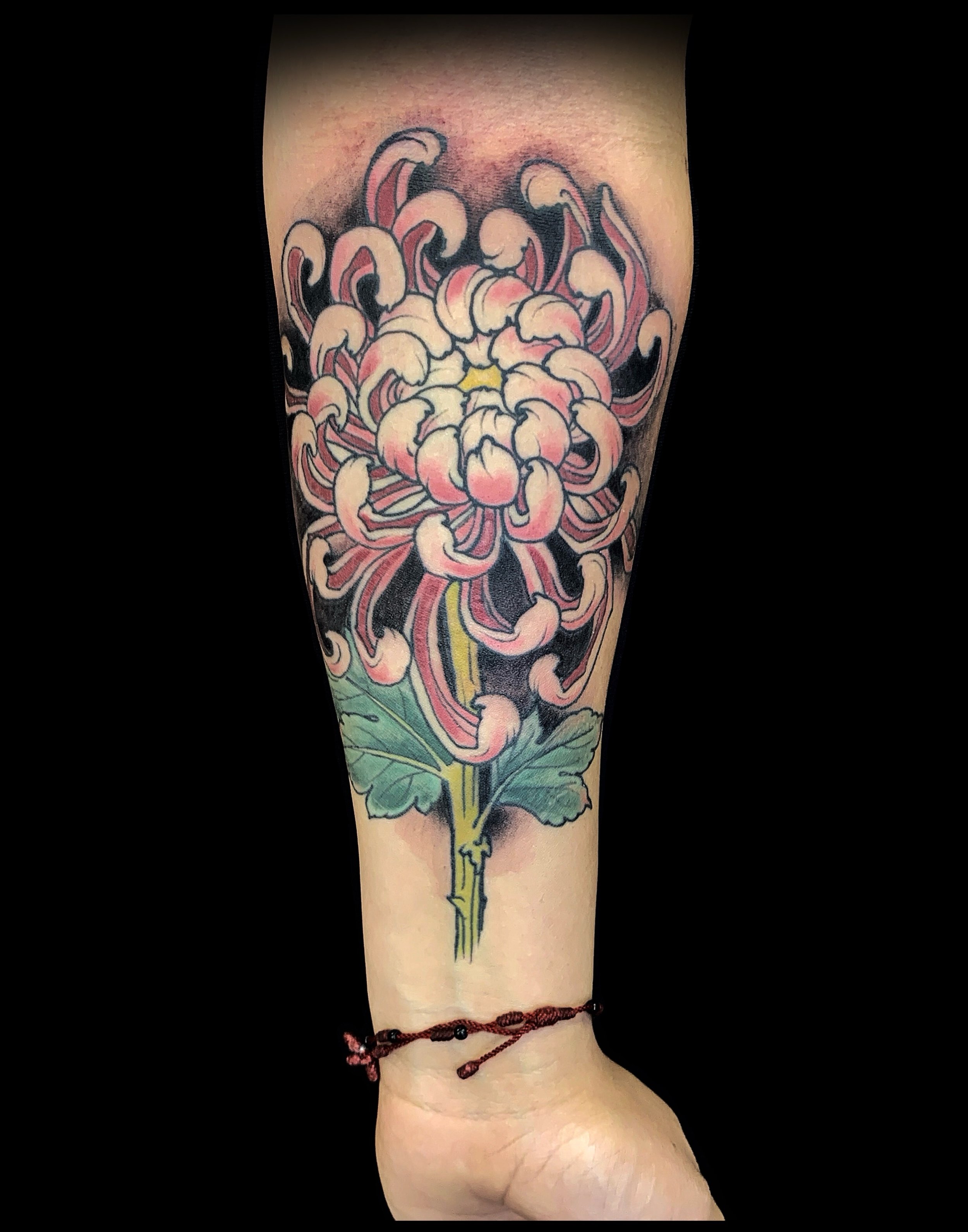 Tattoo of a large pink and white flower with green leaves on a person's forearm, with a maroon string bracelet around the wrist.
