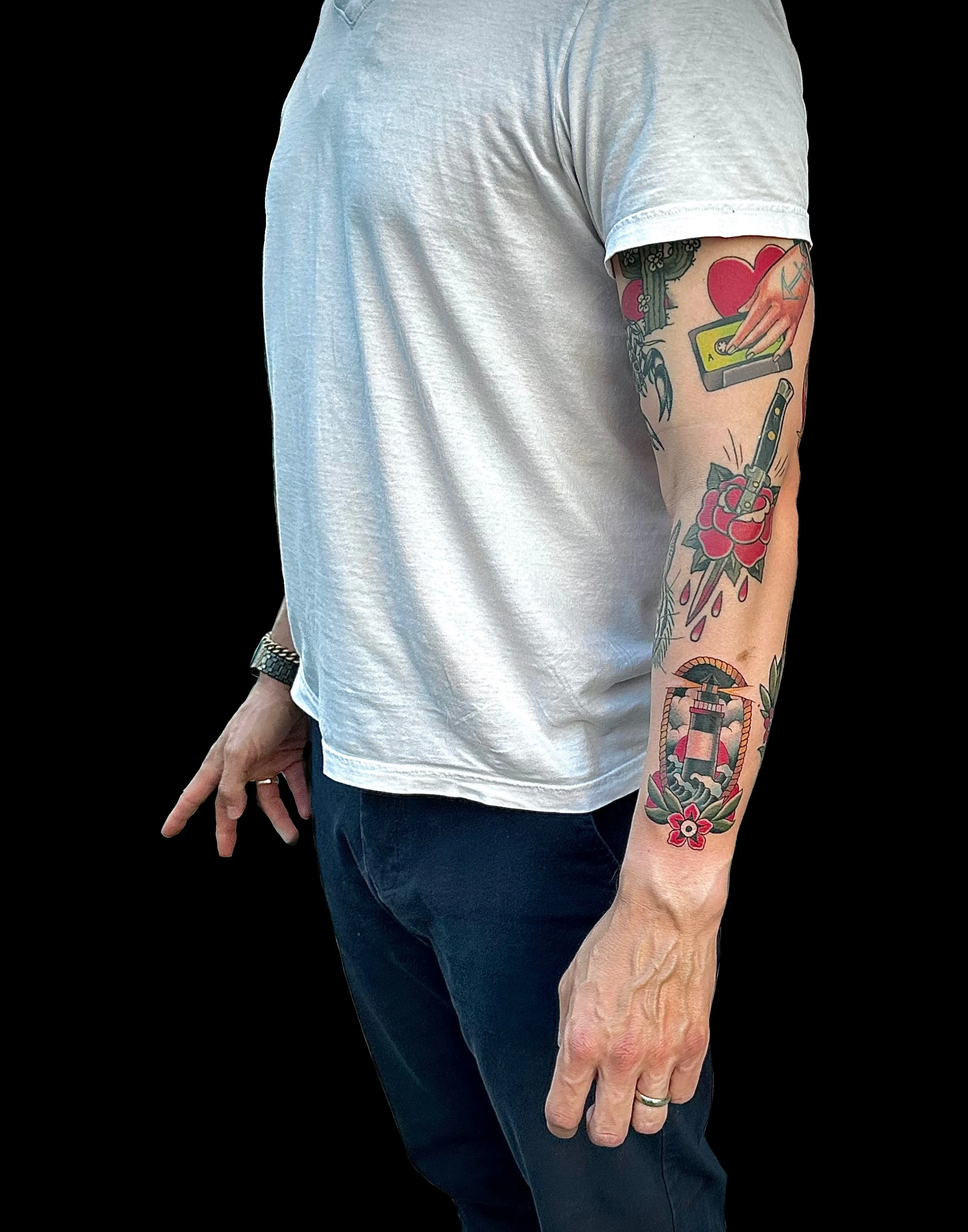 A person wearing a white T-shirt and dark pants, showing multiple colorful tattoos on their right arm, including a heart, a cassette tape, a flower, a knife, and other designs.