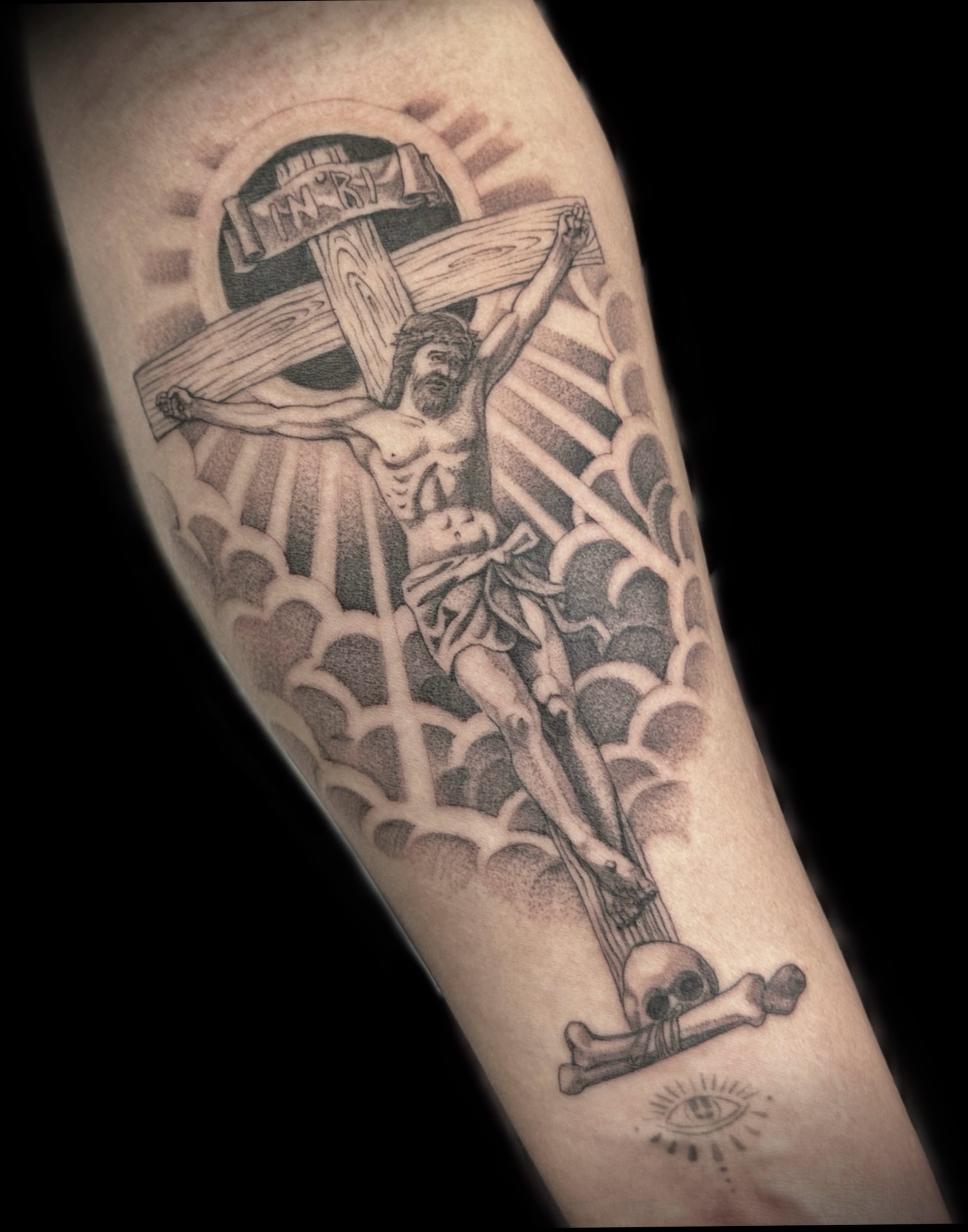 Tattoo of Jesus Christ on the cross with a sunburst background, a banner reading 'INRI', and a skull at the bottom, with additional smaller symbols like an eye and a compass.