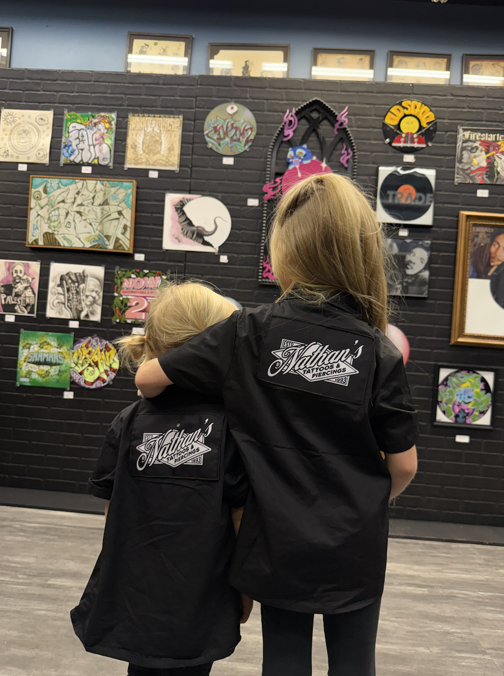 Two young girls with blonde hair, wearing matching black shirts with 'Nathan's Tattoos & Piercings' logos, standing in front of a black brick wall displaying various colorful artwork and paintings.