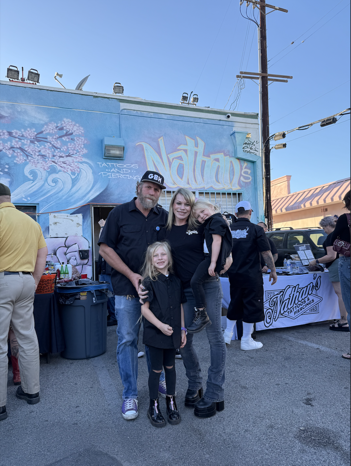 A family of four stands in front of a tattoo shop outdoor event, with people and a booth visible in the background. The family includes a man with a beard, a woman, and two young girls, all smiling.
