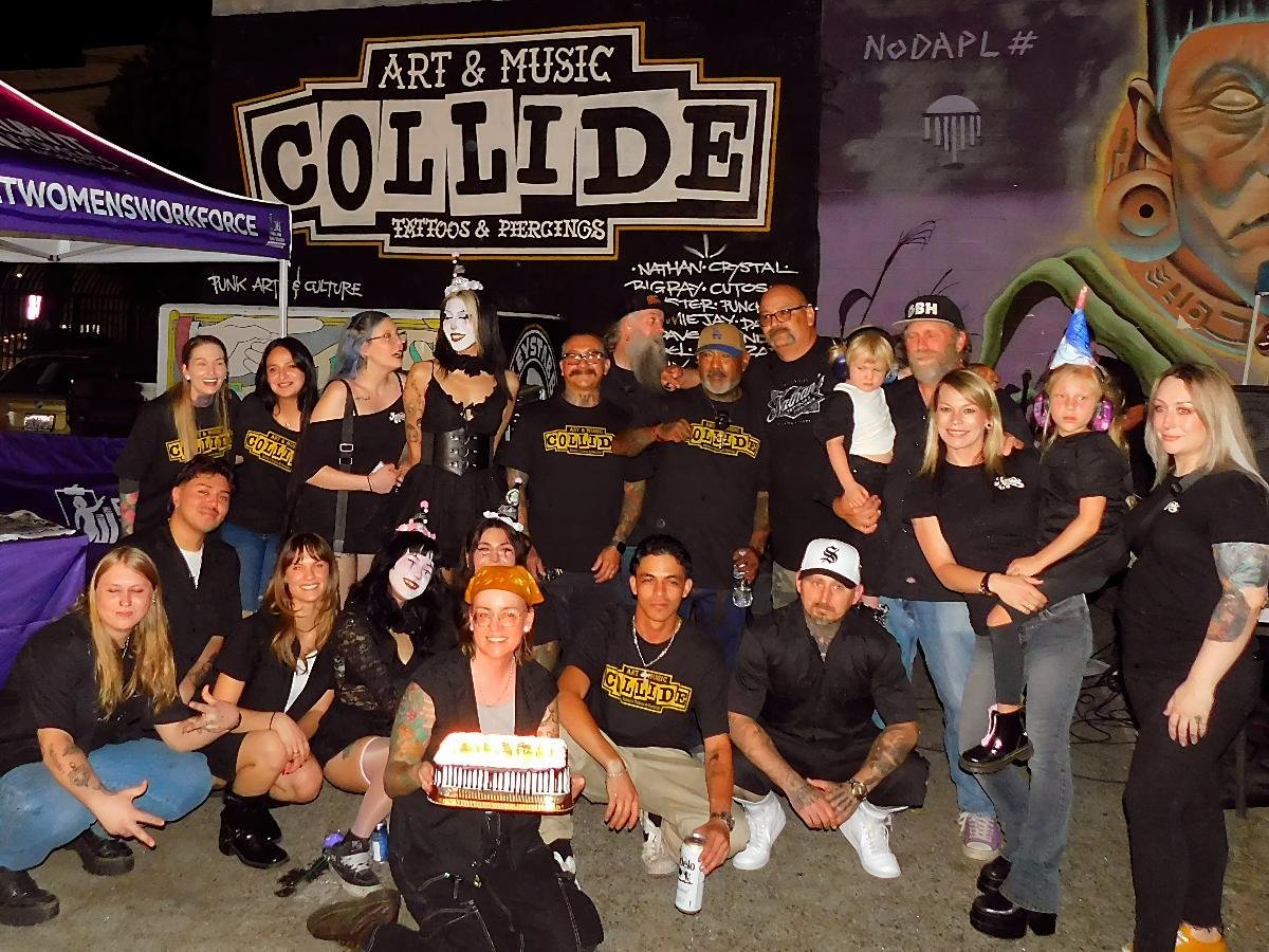 Group of people celebrating in front of a mural and a banner that reads "Art & Music Collision Tattoos & Piercings." Some people are wearing black shirts with the same logo, others are dressed in costumes and face paint, holding a cake and drinks. Th