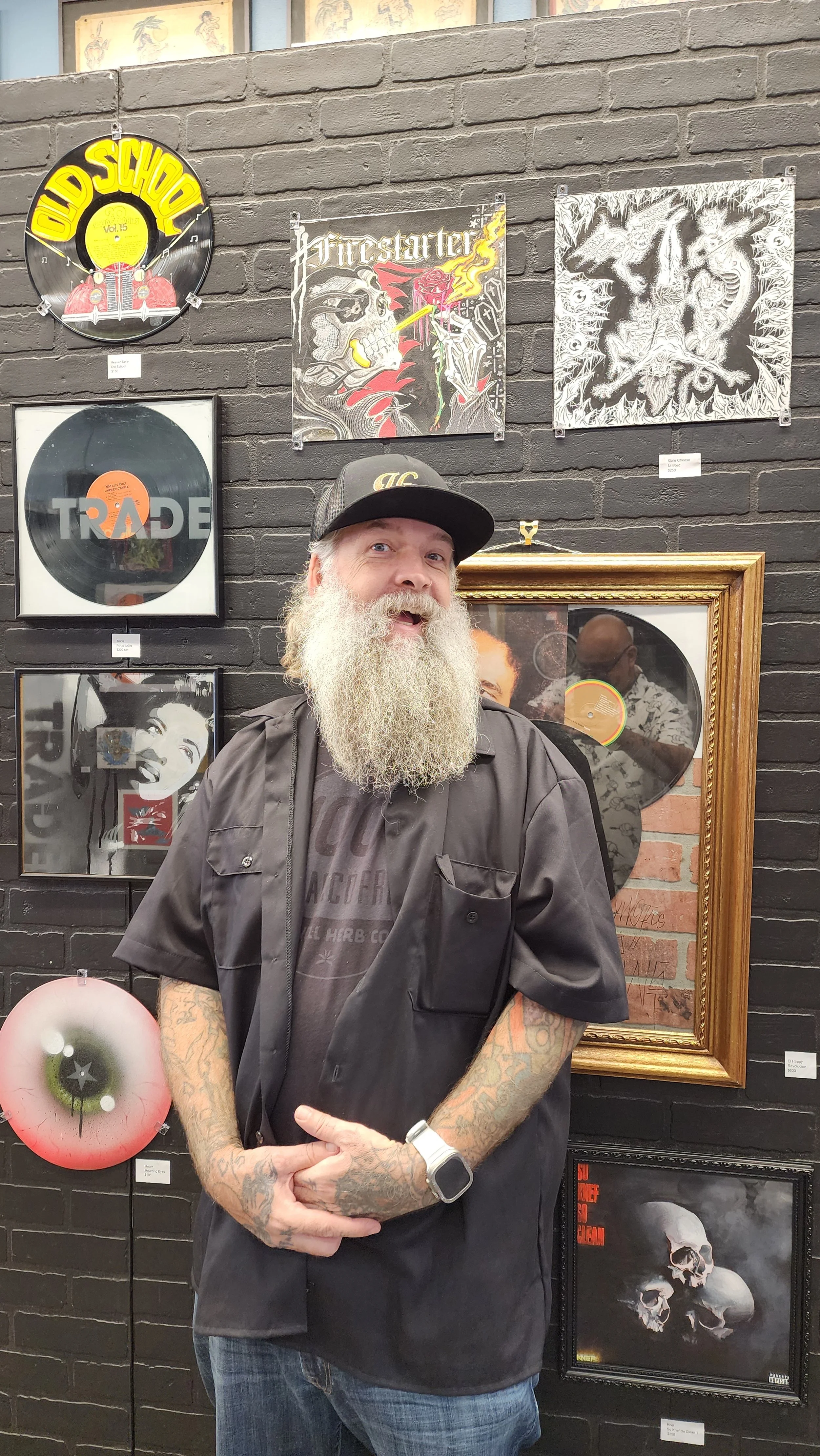 A man with a long white beard, tattoos, and a black cap standing in front of a black brick wall display of framed artwork and vinyl records. The artwork features skulls, a skull smoking a cigarette, a gorilla, and a woman with a skull mask. The vinyl