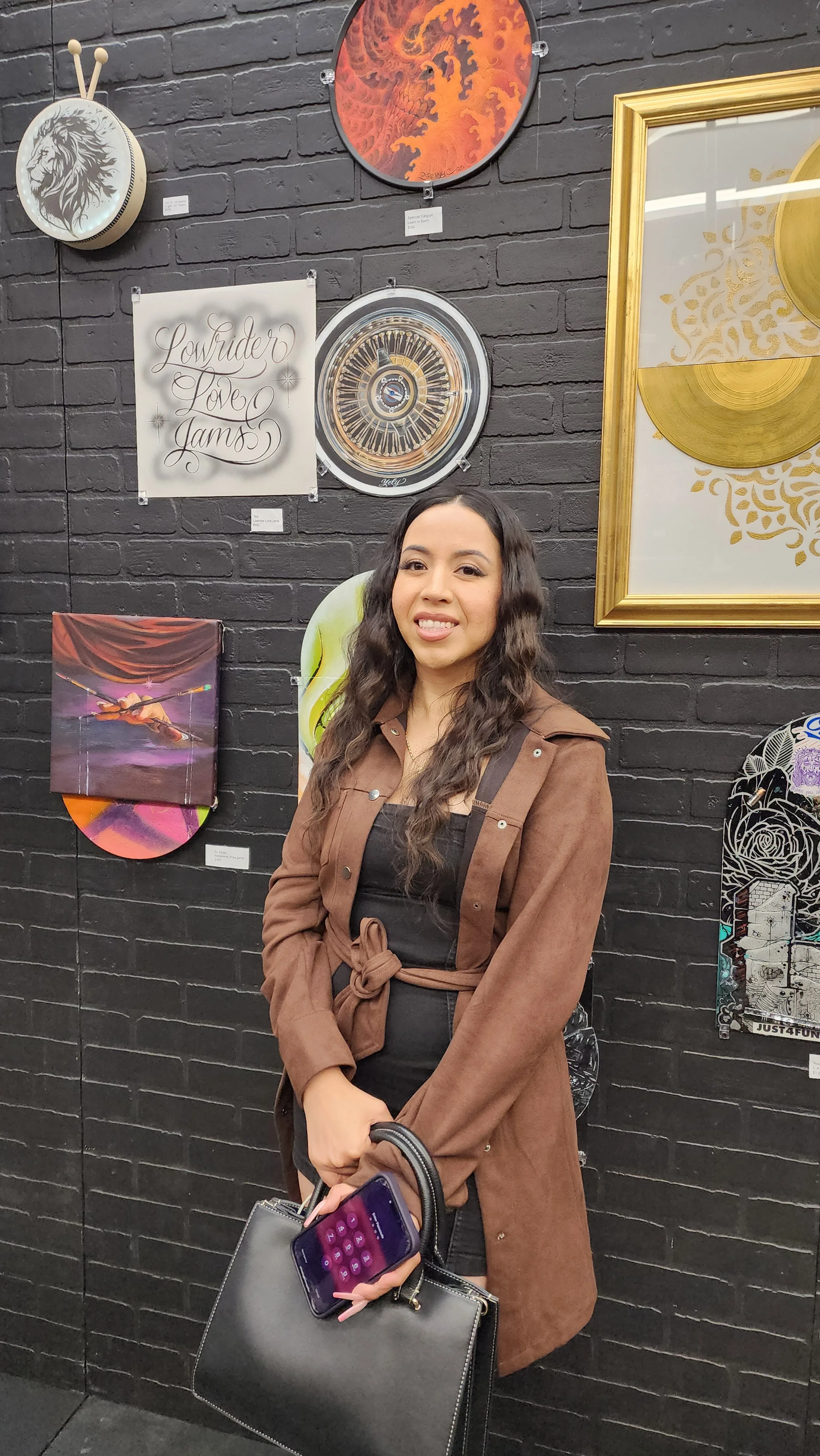 Young woman with long wavy hair, wearing a brown trench coat, smiling at artwork displayed on a black brick wall at an art gallery. She holds a smartphone, a black handbag, and a gray tote bag.