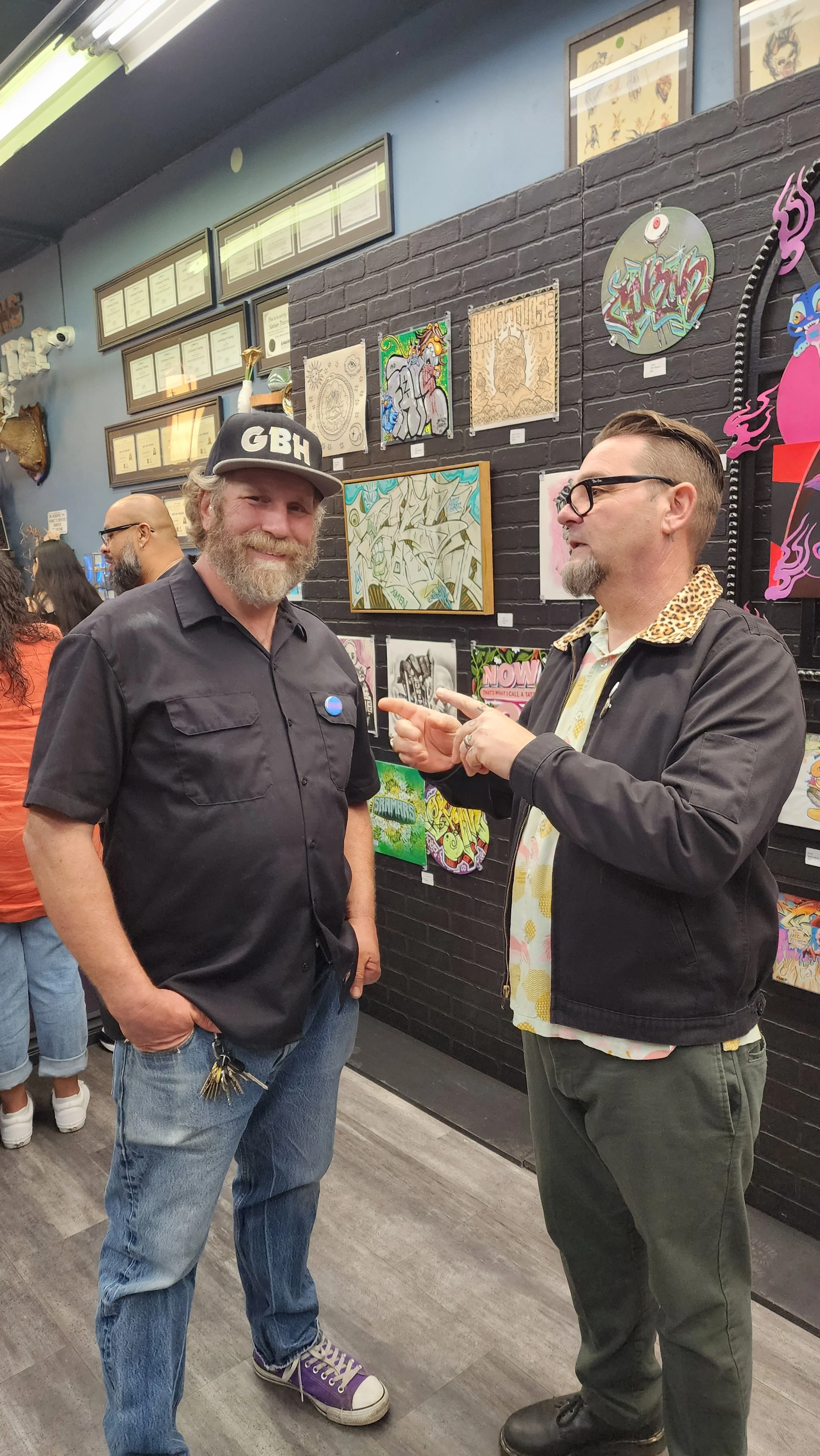 Two men are having a conversation in an art gallery. The man on the left is wearing a black cap with "GBH" on it, a black shirt, and jeans, while the man on the right is wearing glasses, a black jacket with a leopard-print collar, and dark pants. The