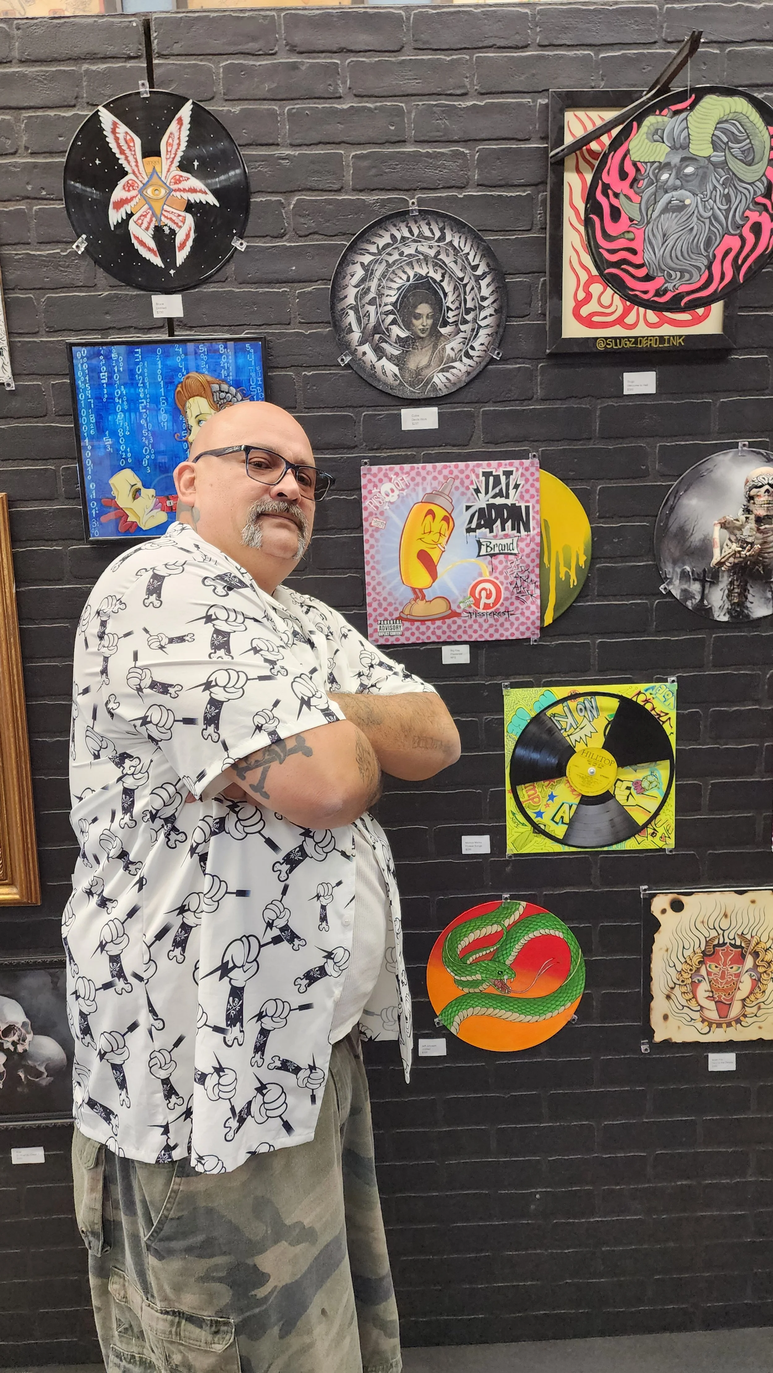 A man with glasses and a mustache, wearing a white shirt with a black cartoon pattern, stands with arms crossed in front of a black brick wall decorated with various artwork. The artwork includes colorful, stylized images like a snake, a person with 