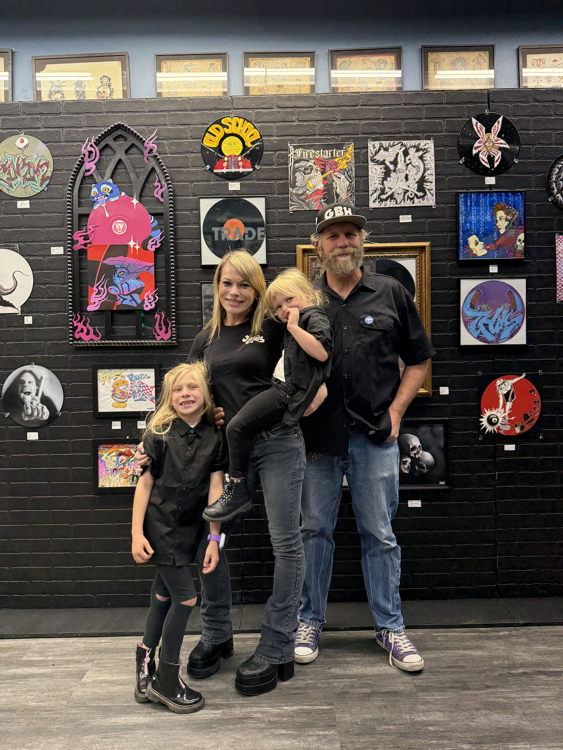 Family of four, two women and two young girls, posing in front of a black brick wall displaying various colorful framed artworks and paintings. The family is smiling and dressed in black, with the women and one girl wearing black shirts and the other