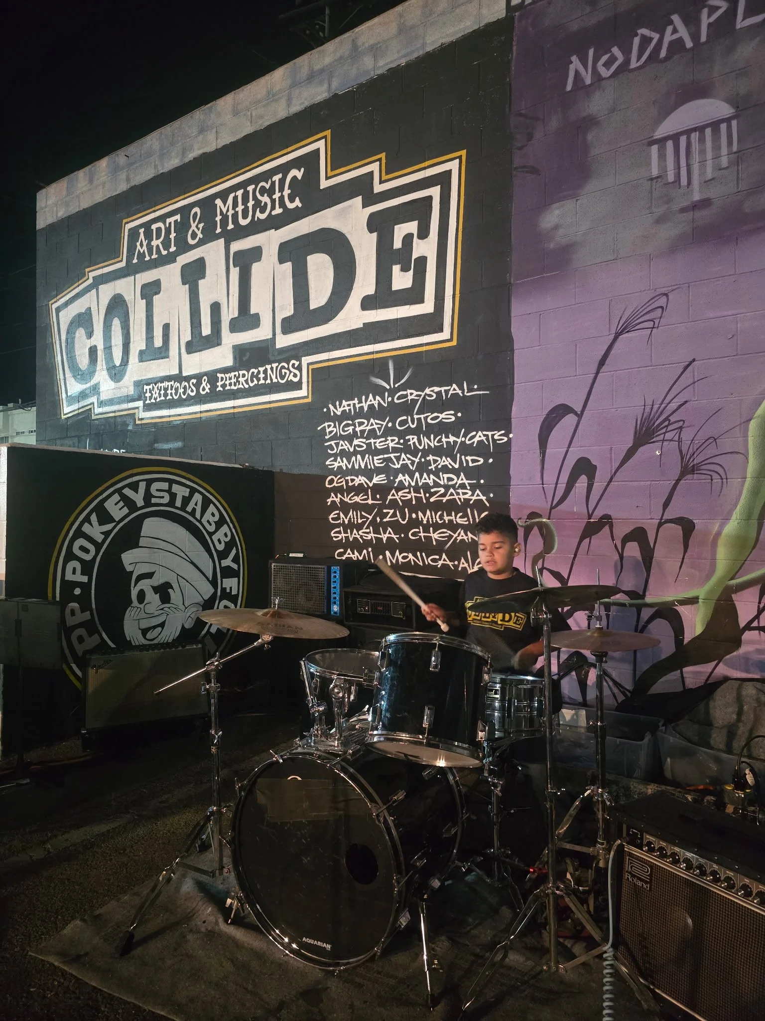 A young boy playing drums at an outdoor event during nighttime, with a large sign in the background reading 'Art & Music Collective Tattoos & Piercings' and a list of names. The scene includes a drum set and an amplifier, with colorful mural art on t