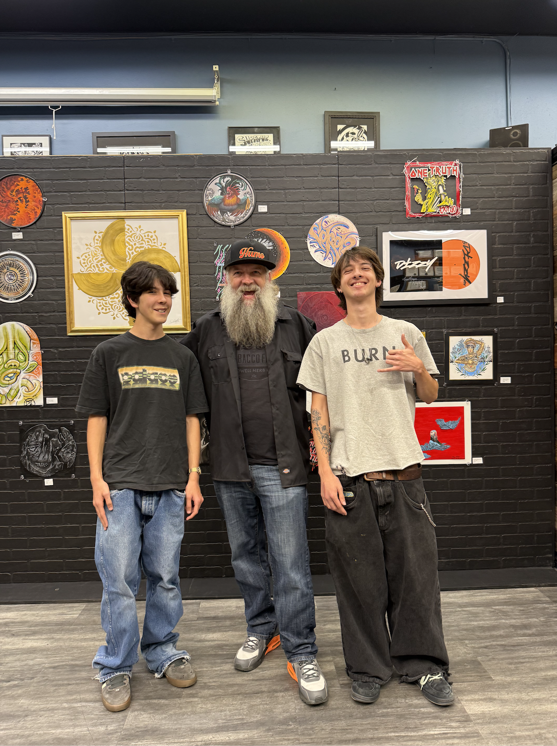 Two young men and an older man with a long beard standing inside an art gallery in front of a black brick wall decorated with various colorful paintings and art pieces.