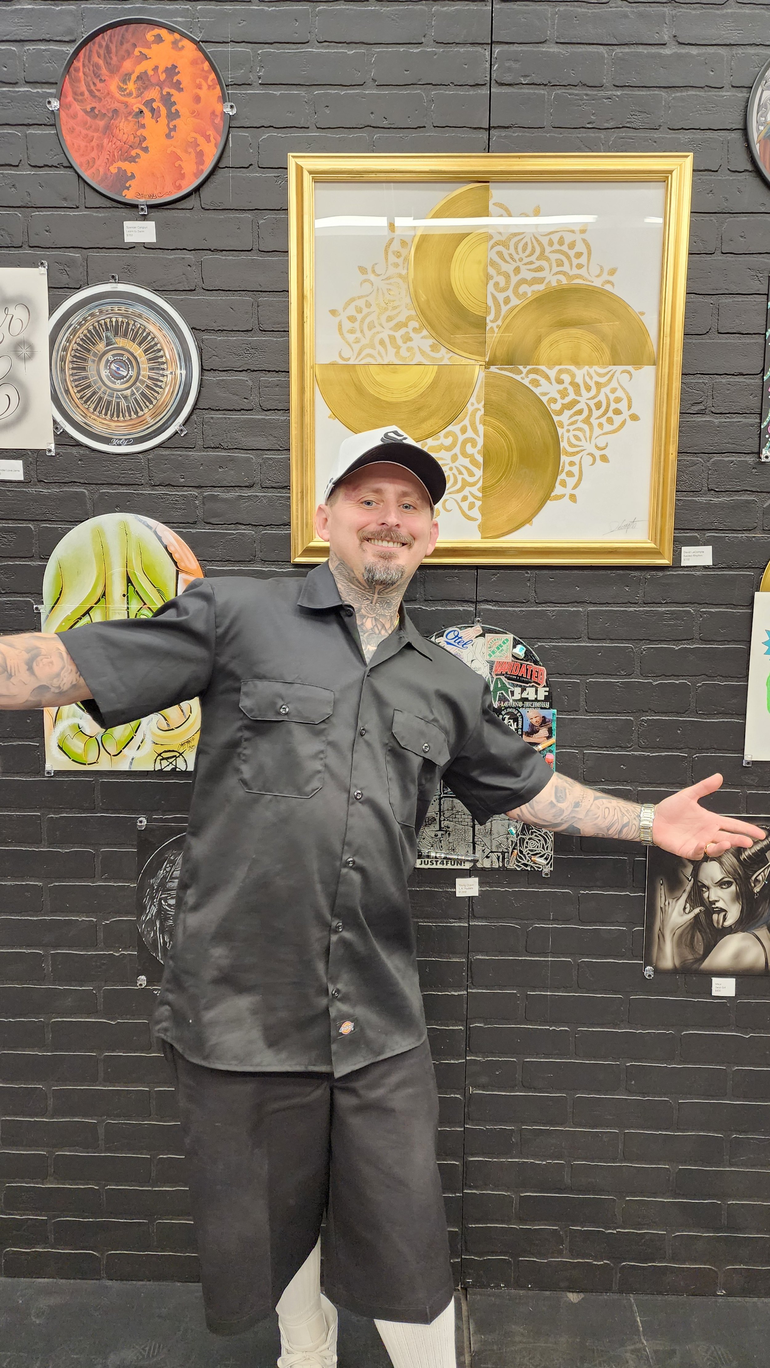 A man with tattoos and a baseball cap smiling with arms open, standing in front of a black brick wall decorated with various art pieces, including a framed gold disc art piece and other paintings and artworks.