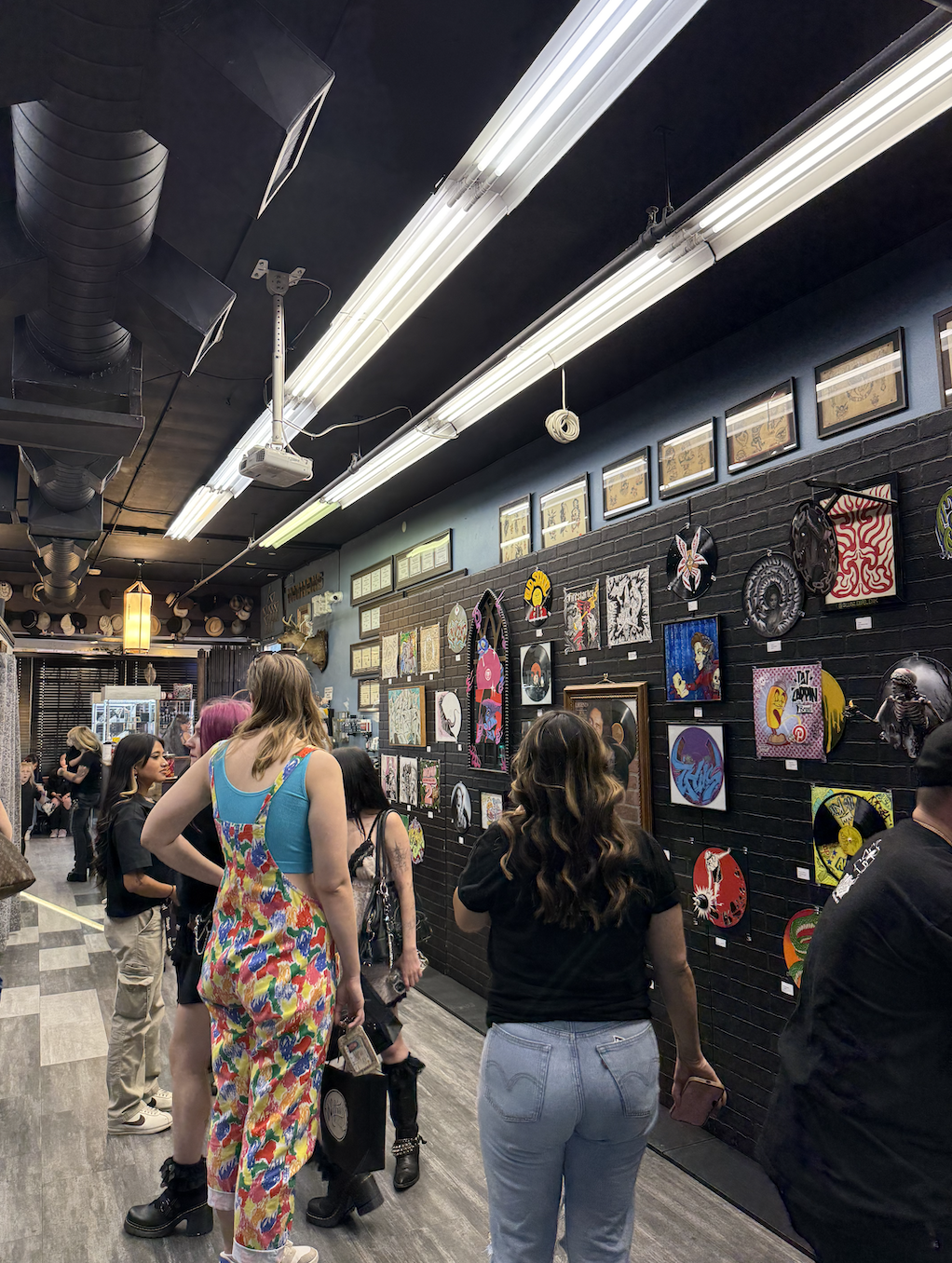 People viewing a wall with framed artwork and vinyl records at an art gallery or exhibit.