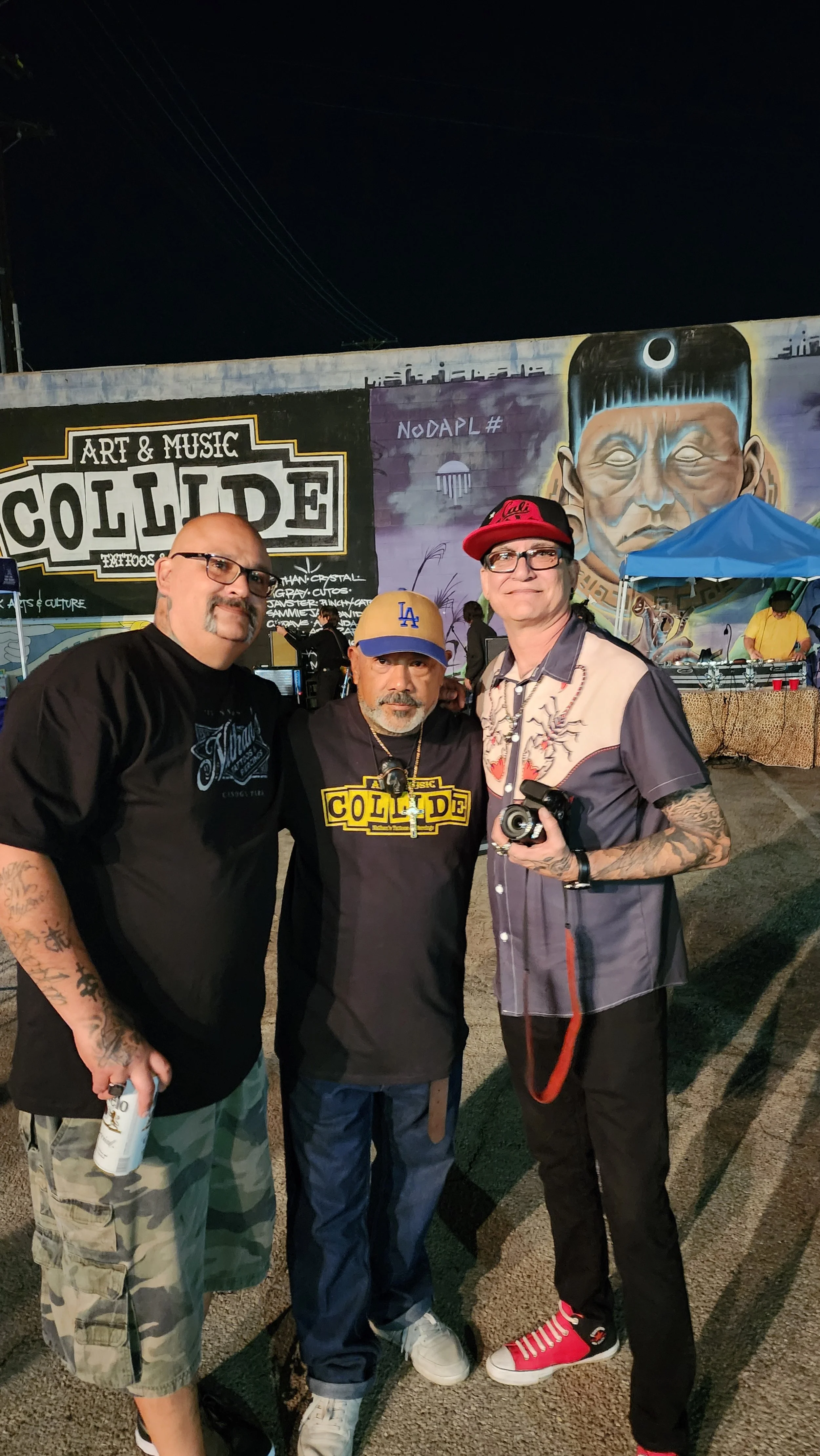 Three men standing together at night in front of a mural that reads 'Art & Music Collide' with a large face painting. One man is holding a camera, and another holds a tube of spray paint. They are at an outdoor event with tents and a DJ in the backgr