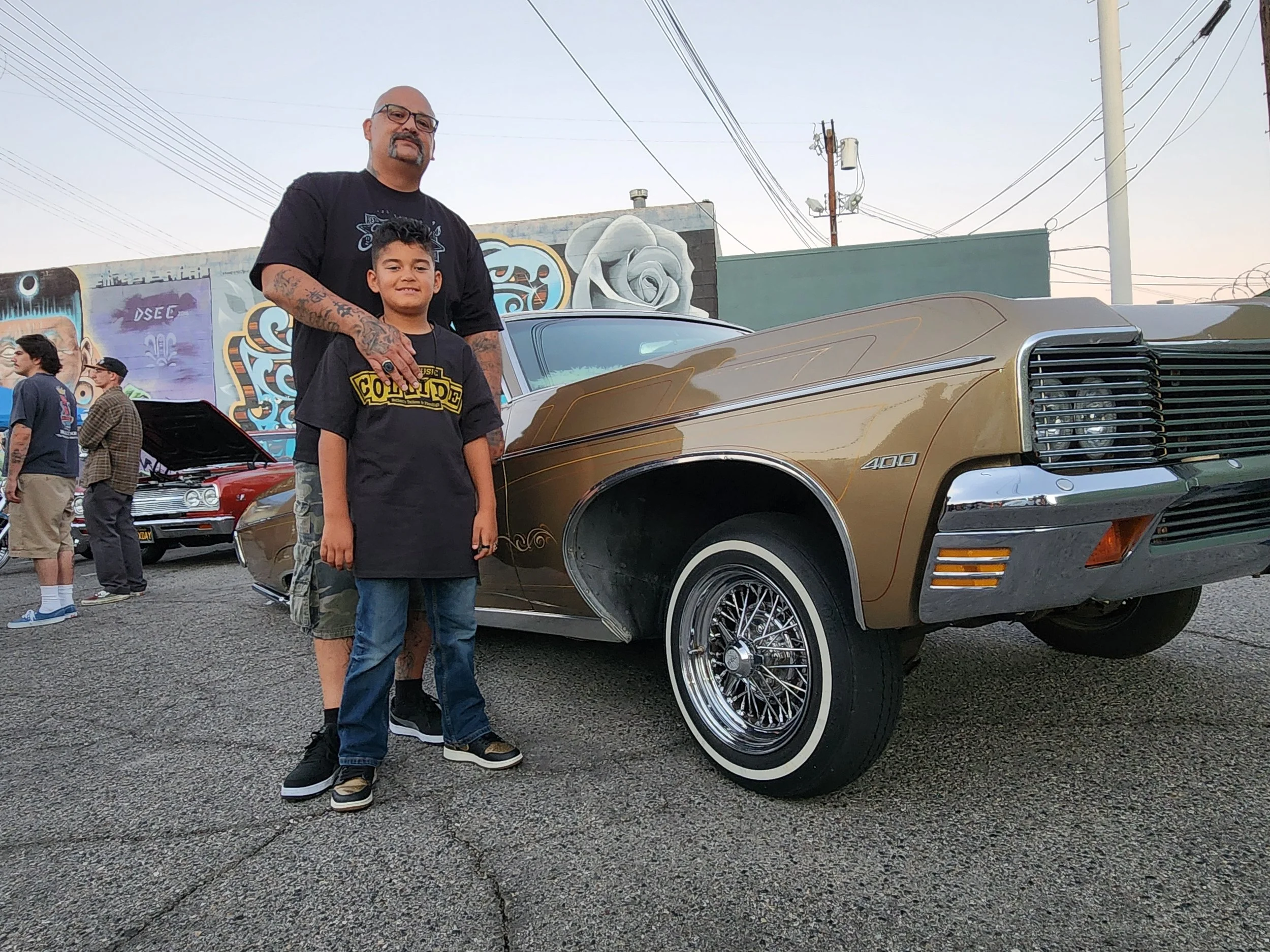 A man and a young boy standing in front of a gold-colored vintage car with wire-spoke wheels at an outdoor car show. There are other people and cars in the background, along with colorful murals on the wall.