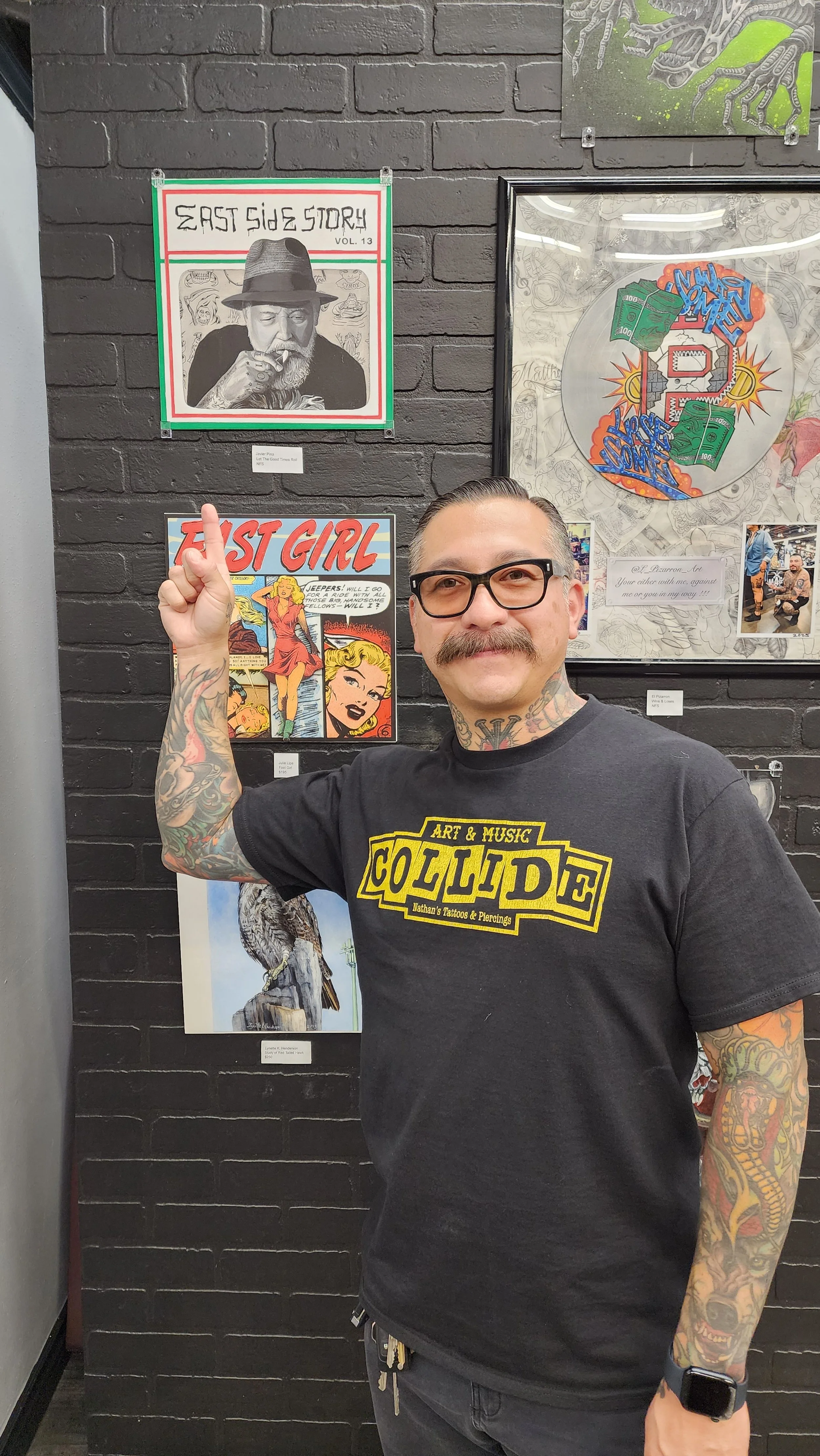 A man with glasses and tattoos pointing up with his right hand, wearing a cuffed black t-shirt with yellow text, standing in front of a black brick wall with various art posters. The posters include a sketch of a man with a hat, a comic book style gi